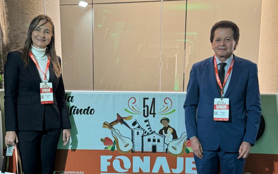 Fotografia colorida que mostra uma mulher e um homem posando pra foto em frente a um banner escrito Fonaje