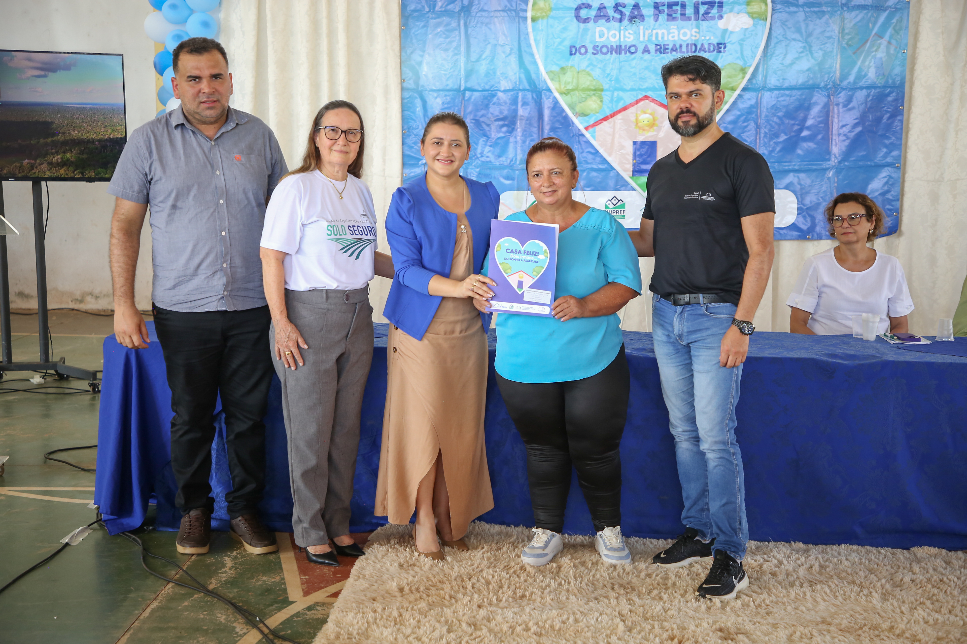 Fotografia colorida que mostra a corregedora geral de Justiça e um juiz posam pra foto com uma moradora segurando o título de terra 