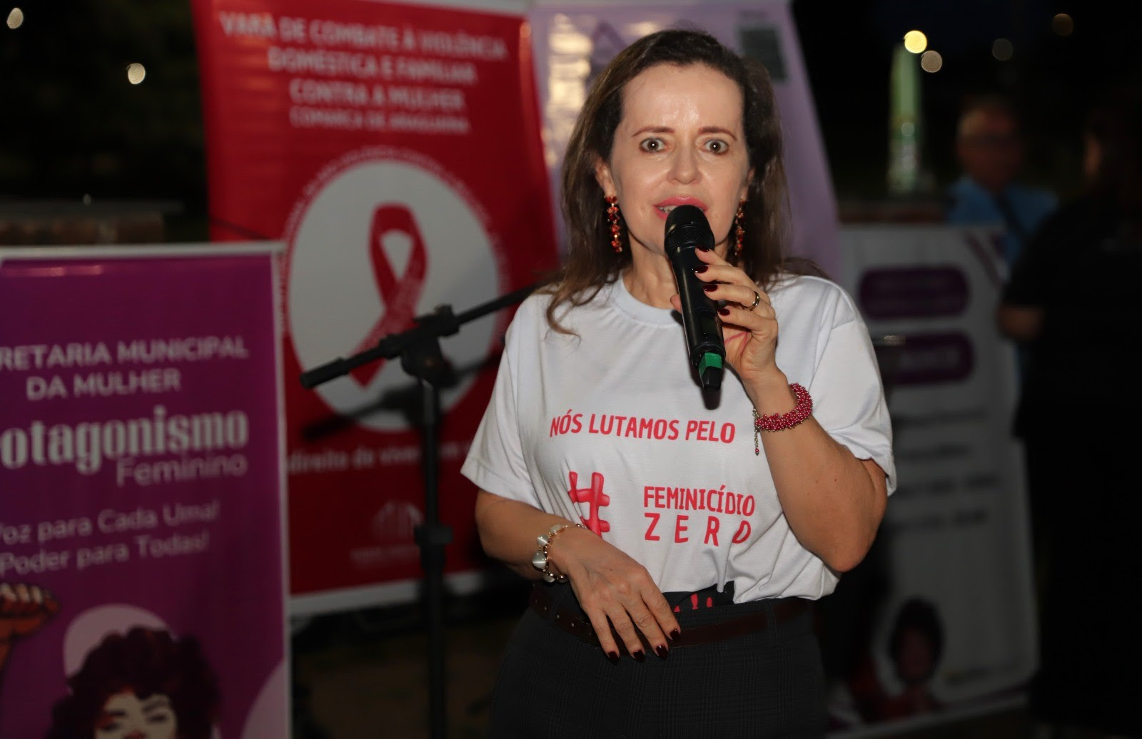 Imagem colorida que mostra uma mulher branca, usando uma camisa branca de ação contra o feminicídio, falando ao microfone