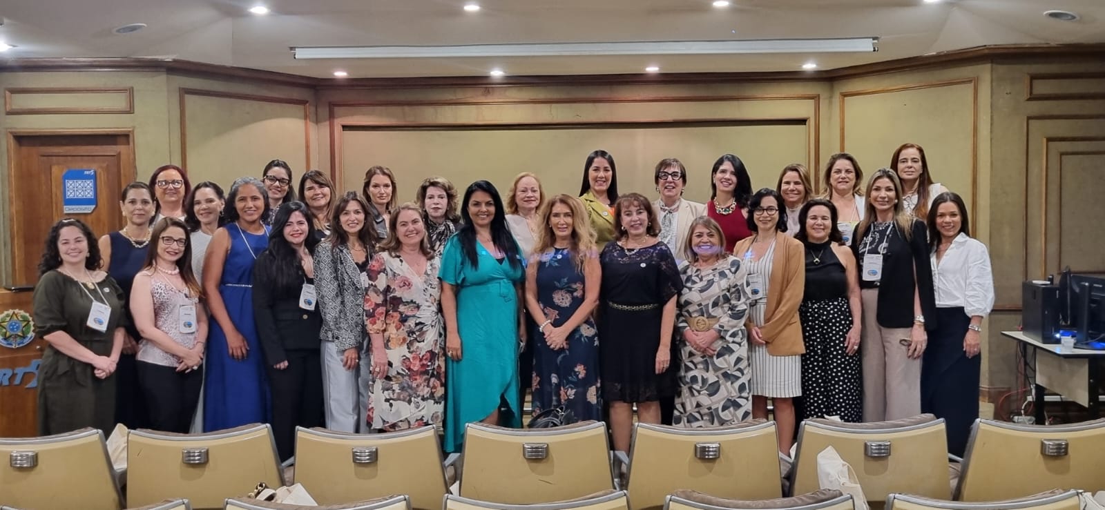 Fotografia colorida que mostra dezenas de mulheres em pé, posando para a imagem em uma sala fechada. Elas sorriem para câmera