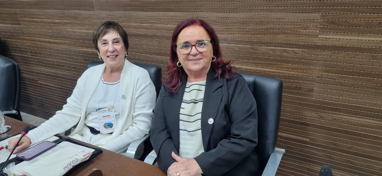 Imagem colorida que mostra duas mulheres brancas (uma de cabelos curtos e outra de cabelos longos), vestindo roupa social e sorrindo para a foto.