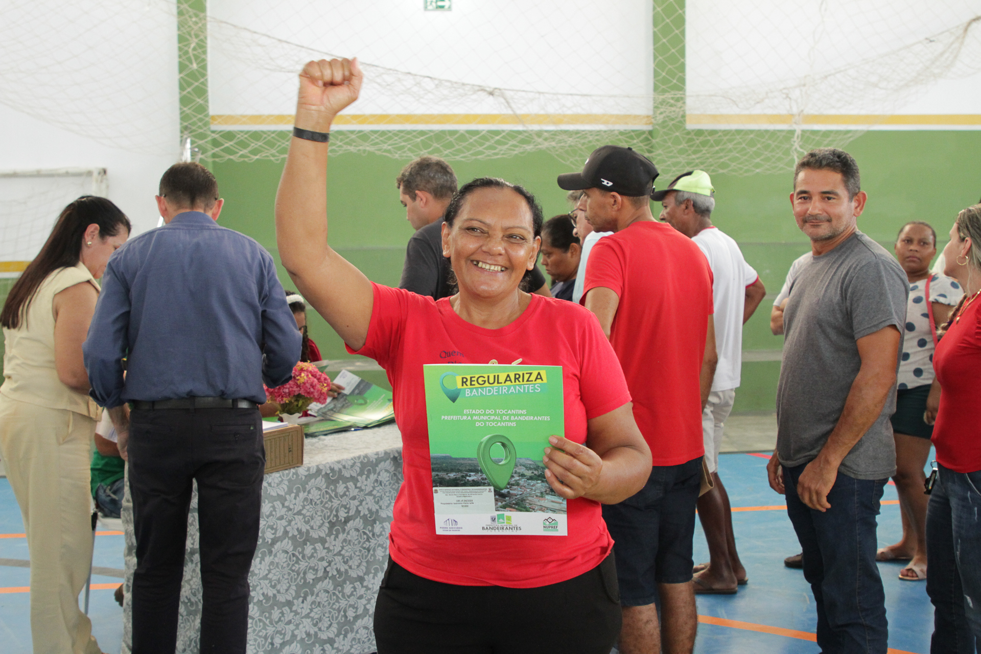 Fotografia colorida que mostra uma mulher negra, de cabelos presos e usando preto e vermelho, com um documento na mão esquerda e com a outra mão levantada com o punho fechado