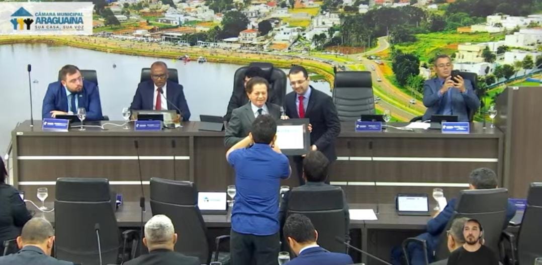 juiz Deusamar recebe certificado das mãos de um vereador; ao fundo aparece a tribuna com outros parlamentares e um painel enorme com a imagem da cidade; à frente parte do auditórico com pessoas assistindo 