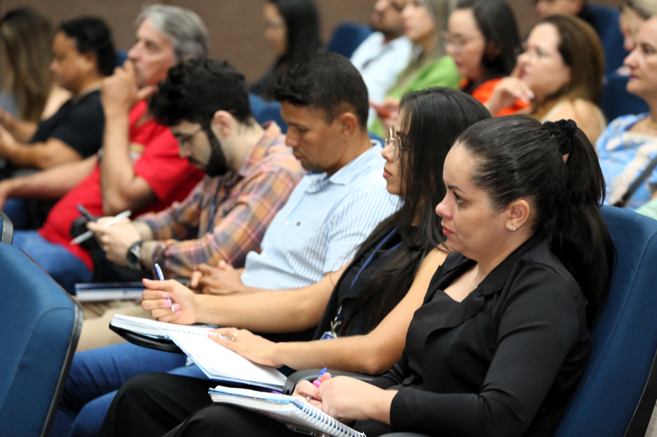 Imagem colorida que mostra pessoas (homens e mulheres) sentadas prestando atenção em uma palestra