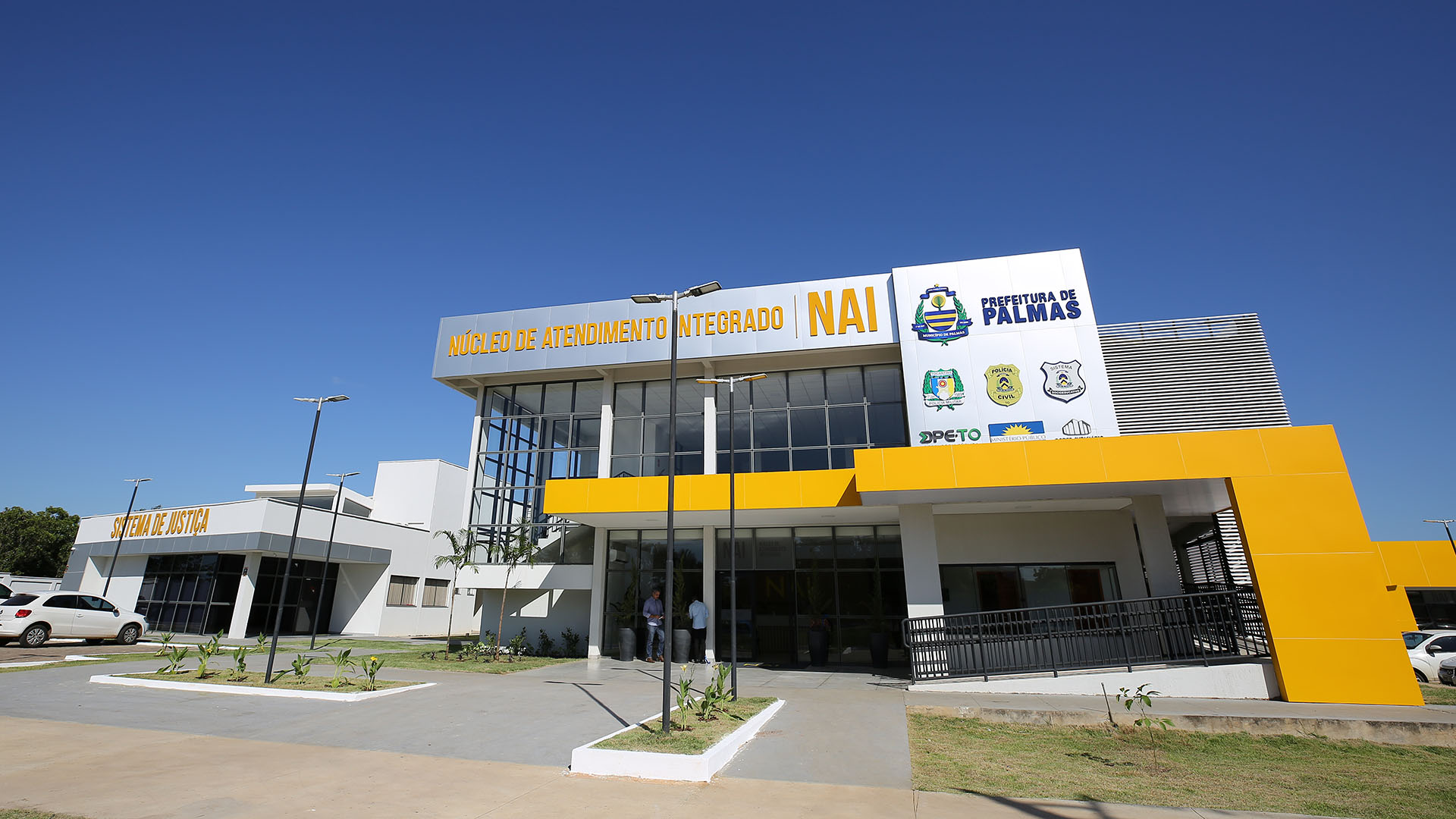 Imagem mostra a fachada do NAI; prédio com fachada de vidro, pintado de amarelo, jardim na frente, rampa de acessibilidade