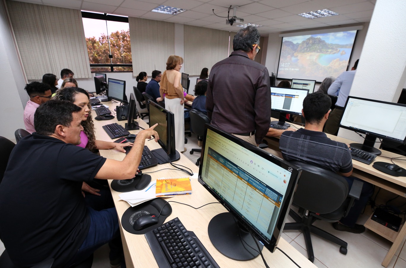 Fotografia colorida de uma sala onde um grupo de quinze pessoas estão mexendo em computadores enquanto são orientadas por pessoas que estão de pé