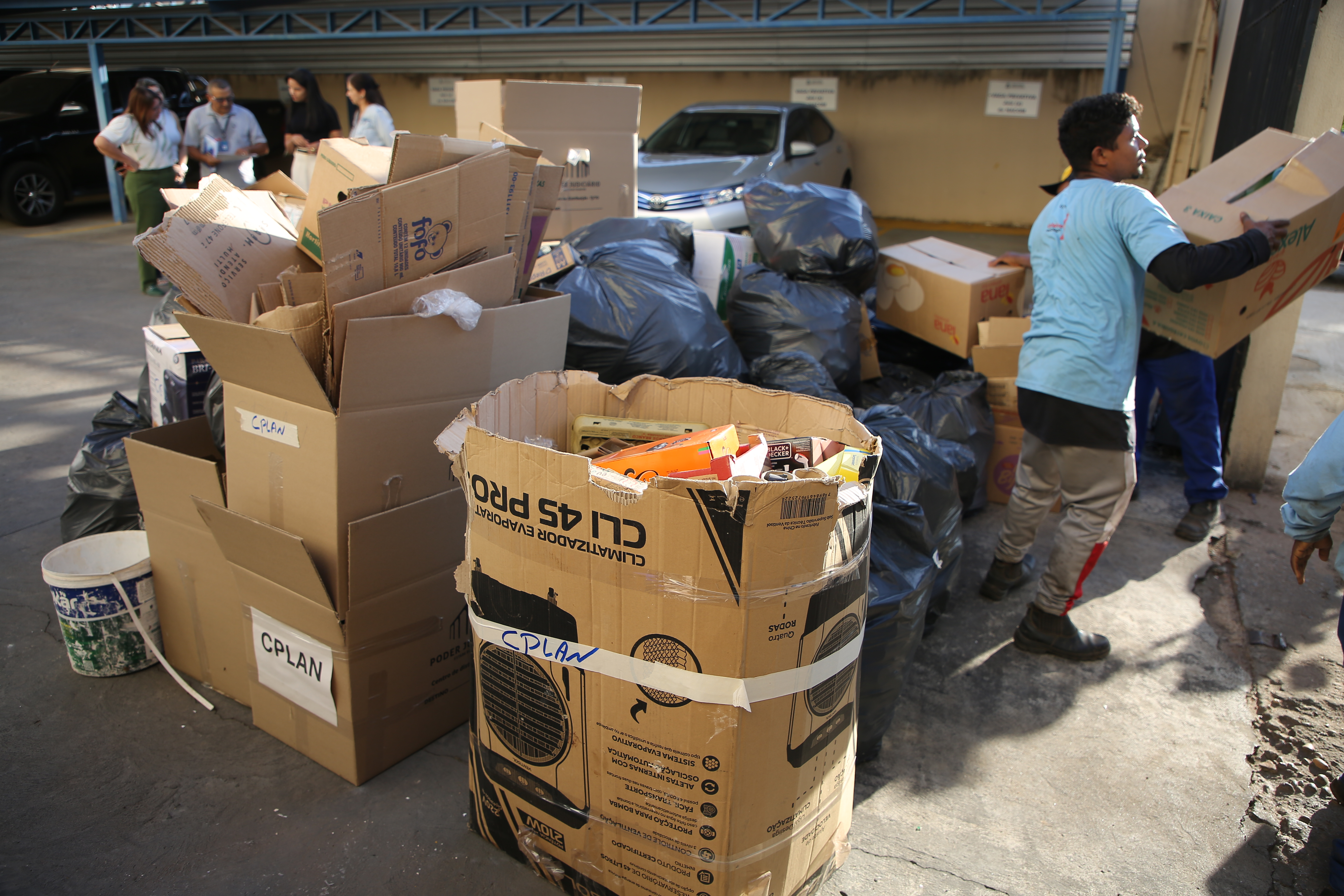 Fotografia colorida com várias caixas e sacos pretos com lixos recicláveis sendo carregados por um homem, e ao fundo quatro pessoas conversando 