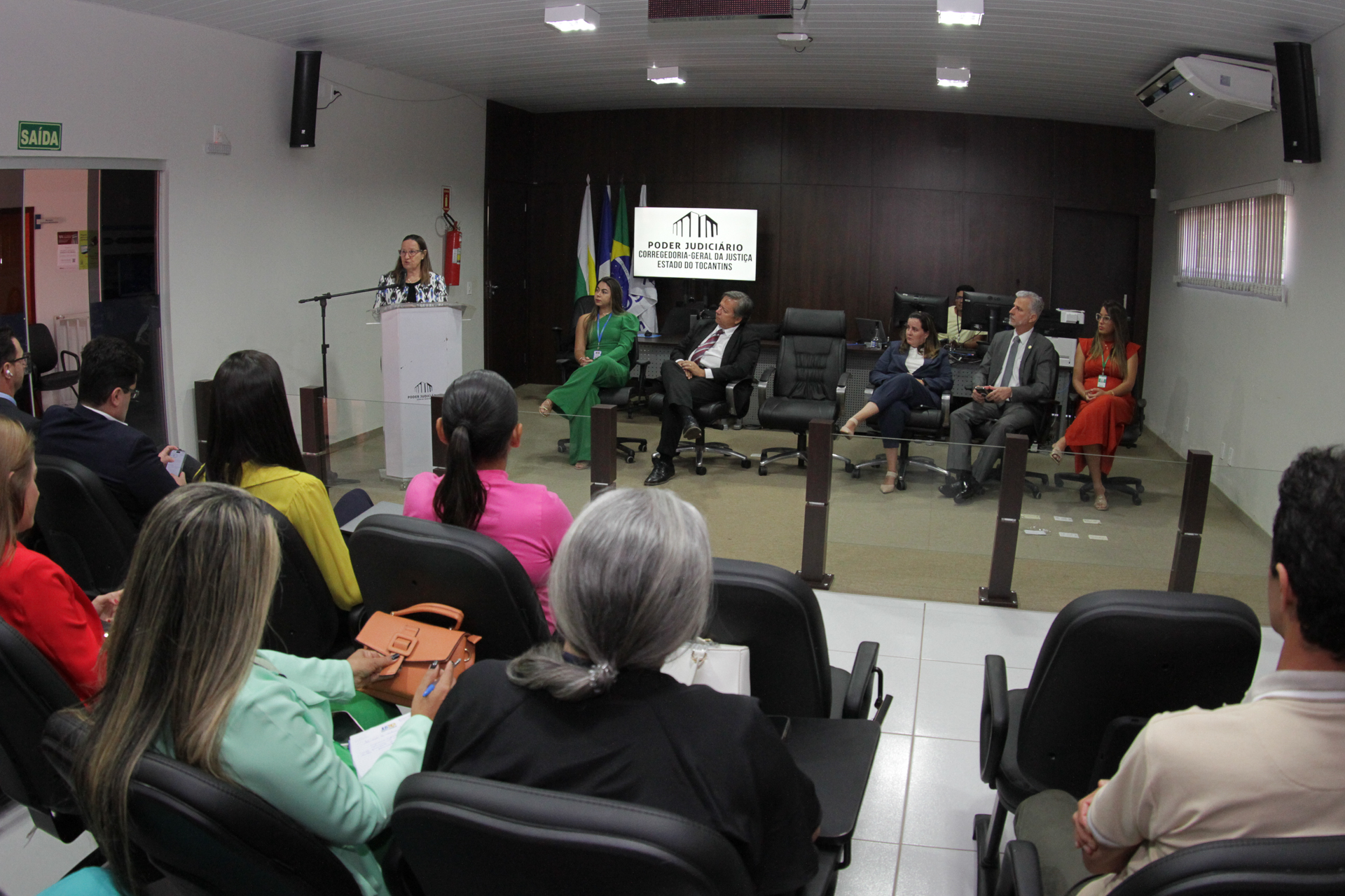 Fotografia colorida onde pessoas estão sentadas em cadeiras pretas em um auditório enquanto cinco pessoas estão sentadas a frente, viradas para eles, e uma mulher de camisa branca com detalhes pretos e cabelo solto está de pé atrás de um palanque branco falando em um microfone. Eles estão em uma sala branca com uma parede de madeira escura no fundo