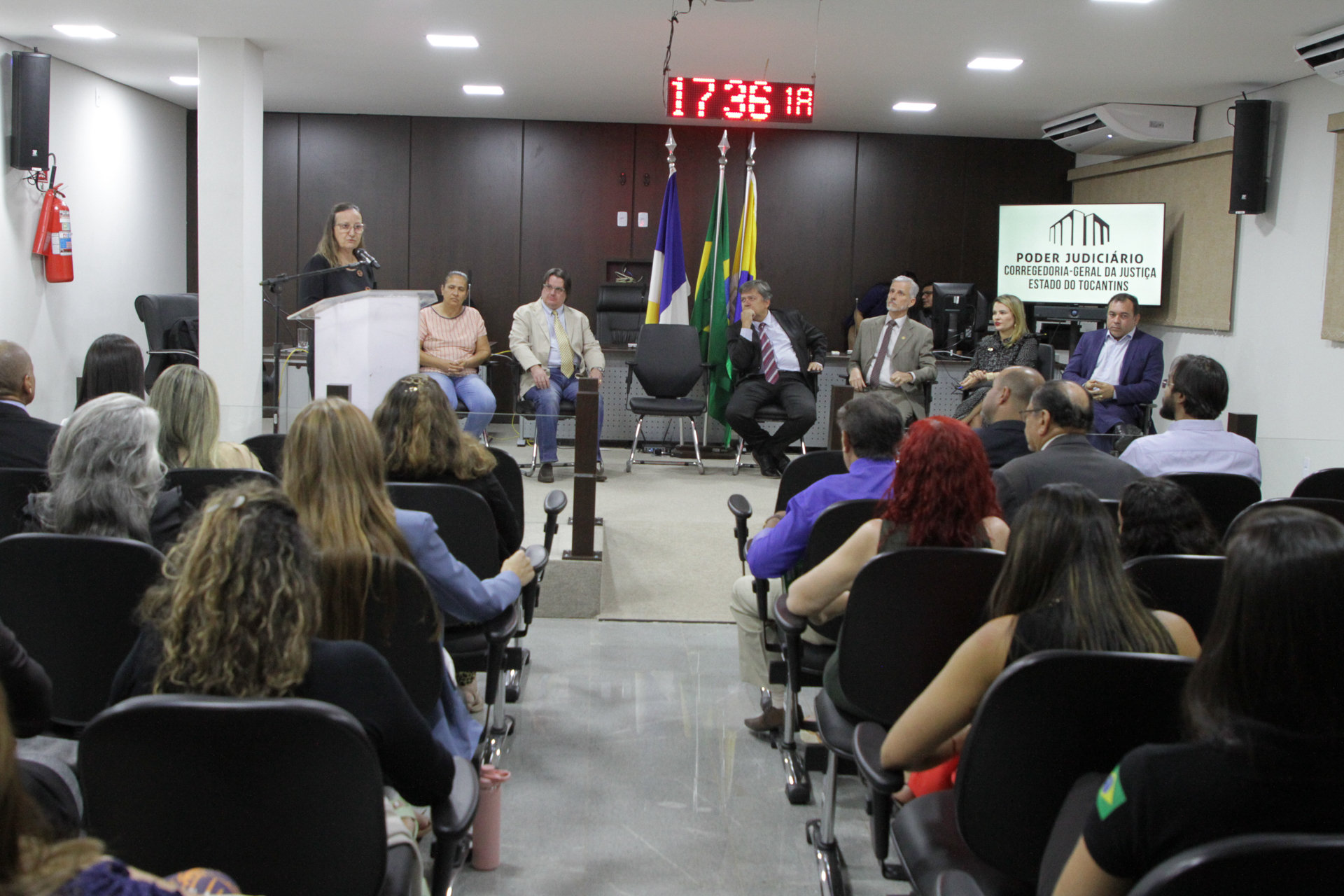 Fotografia colorida onde um grupo de pessoas está sentada em cadeiras de auditório, com uma passarela no meio deles, que leva até um espaço aberto onde quatro homens e duas mulheres estão sentado de frente para eles, em cadeiras pretas, e uma mulher de roupa preta e cabelos claros soltos fala em um microfone atrás de um palanque 