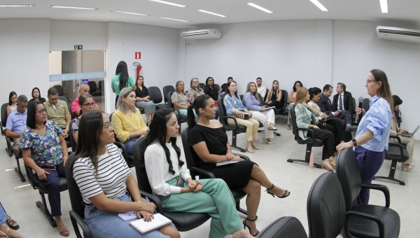 Grupo de pessoas sentadas em cadeiras pretas organizadas em filas olhando para a oradora que está na frente da sala.