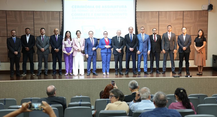 A imagem mostra um grupo de pessoas alinhadas lado a lado em um palco, posando para uma foto. Atrás deles, há uma tela de projeção que exibe o título "Cerimônia de Assinatura de Termo de Cooperação para Combate e Enfrentamento à Desinformação nas Eleições 2024".