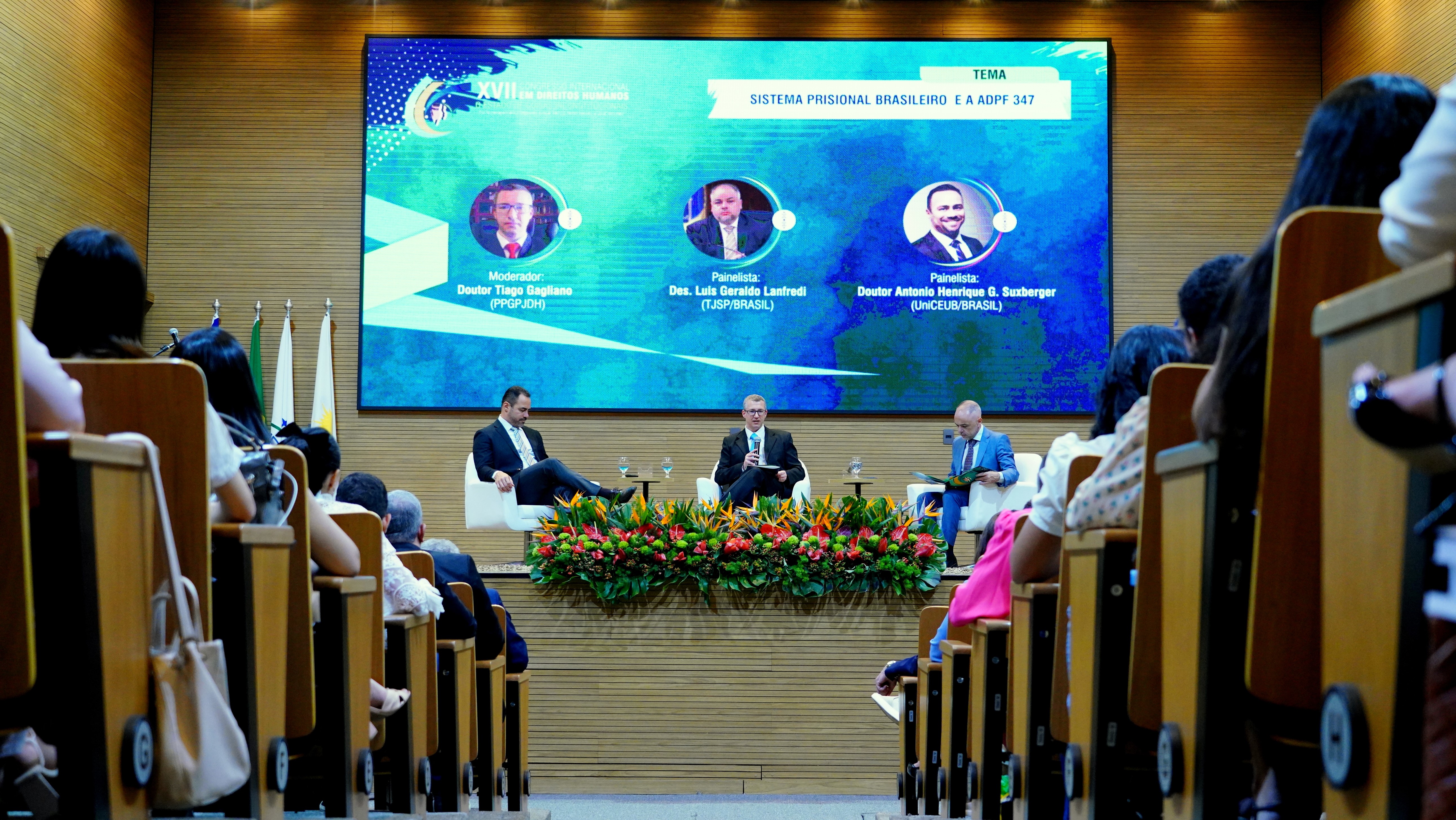Fotografia colorida onde a câmera está no meio de um corredor, e dos dois lados estão pessoas sentadas em cadeiras. Na frente, está um palco com uma tela de fundo e três homens sentados na frente. Eles estão em um auditório coberto de madeira clara 