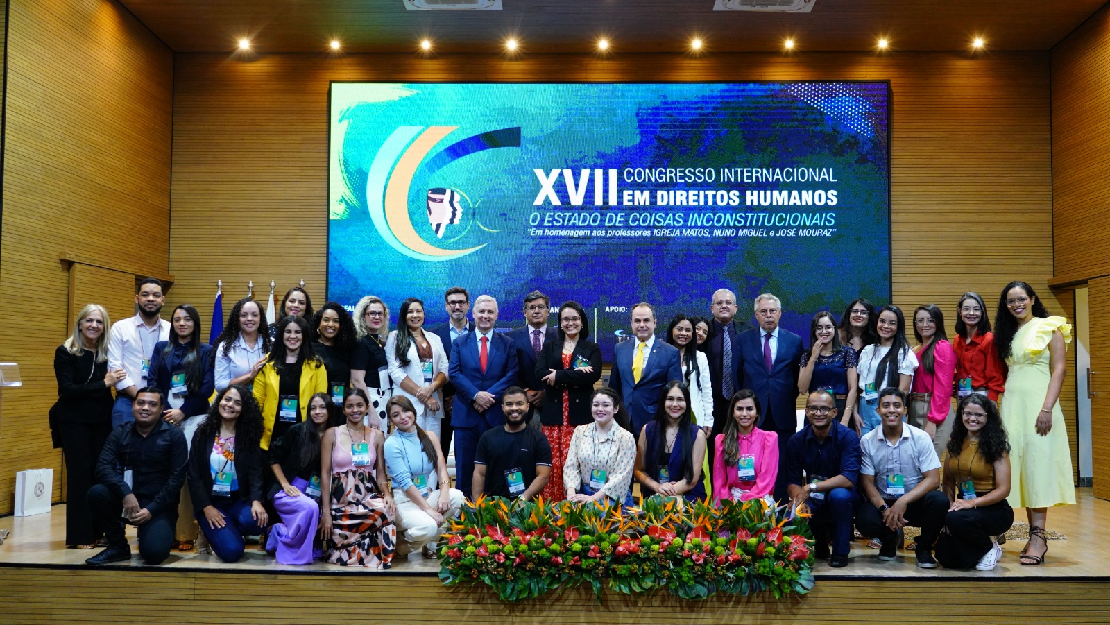 Fotografia colorida onde um grupo de várias pessoas está posando para foto em cima de um palco de madeira clara e um telão azul ao fundo