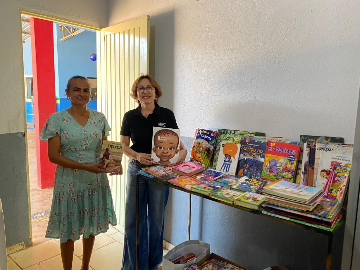 Fotografia colorida de duas mulheres segurando um livro cada uma ao lado de uma mesa cheia de livros. Elas estão paradas em frente a uma porta aberta dentro de uma sala 