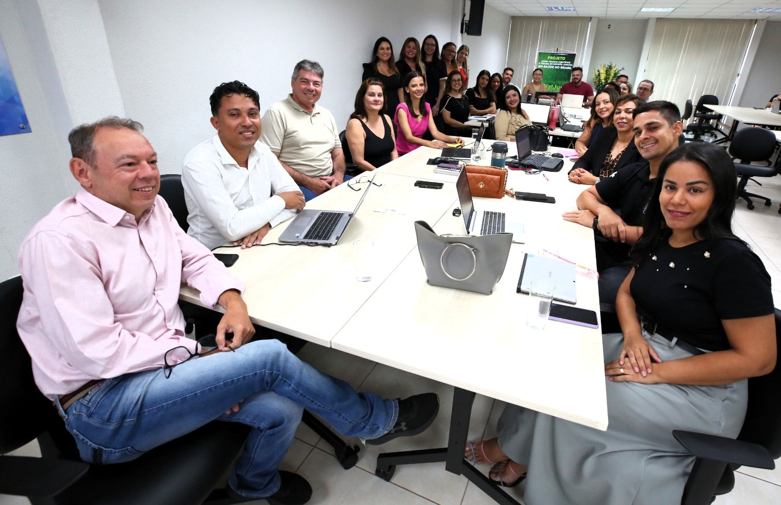 Fotografia colorida de vinte e três pessoas em volta de uma comprida mesa retangular branca com alguns notebooks em cima. A maioria está sentados, e alguns de pé, e eles estão posando para a foto dentro de uma sala branca 