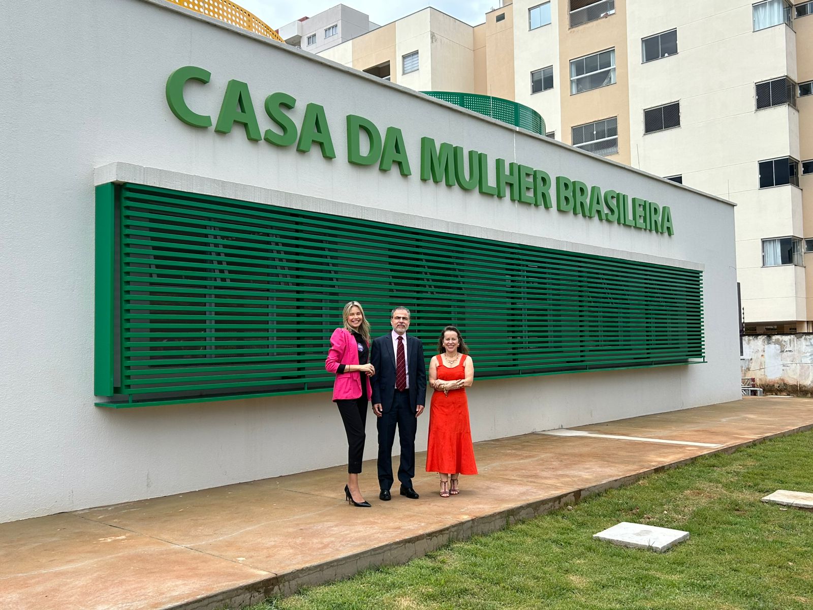 Fotografia colorida onde um homem de terno, uma mulher de vestido vermelho e outra de calça preta e blazer rosa estão parados na frente de um prédio branco e verde, onde uma grande janela de grades verdes se projeta na parede e acima da janela os dizeres "Casa da Mulher Brasileira"