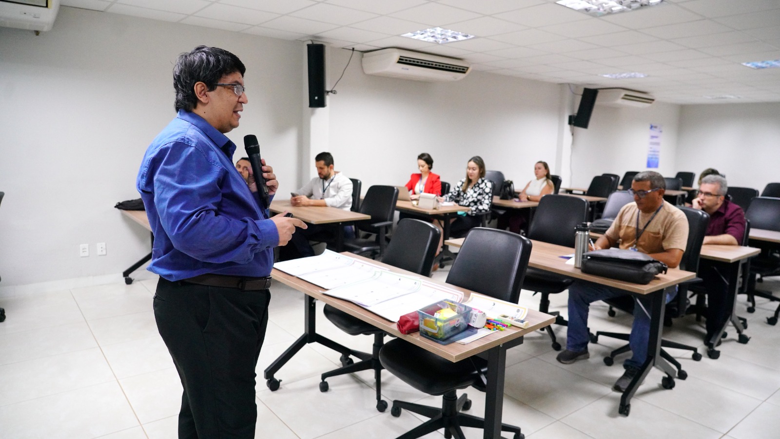 Fotografia colorida onde um homem de camisa azul, calça preta, cabelo preto e óculos segura um microfone enquanto ministra um curso para algumas pessoas que estão sentadas atrás de mesas em uma sala branca com ar condicionados 