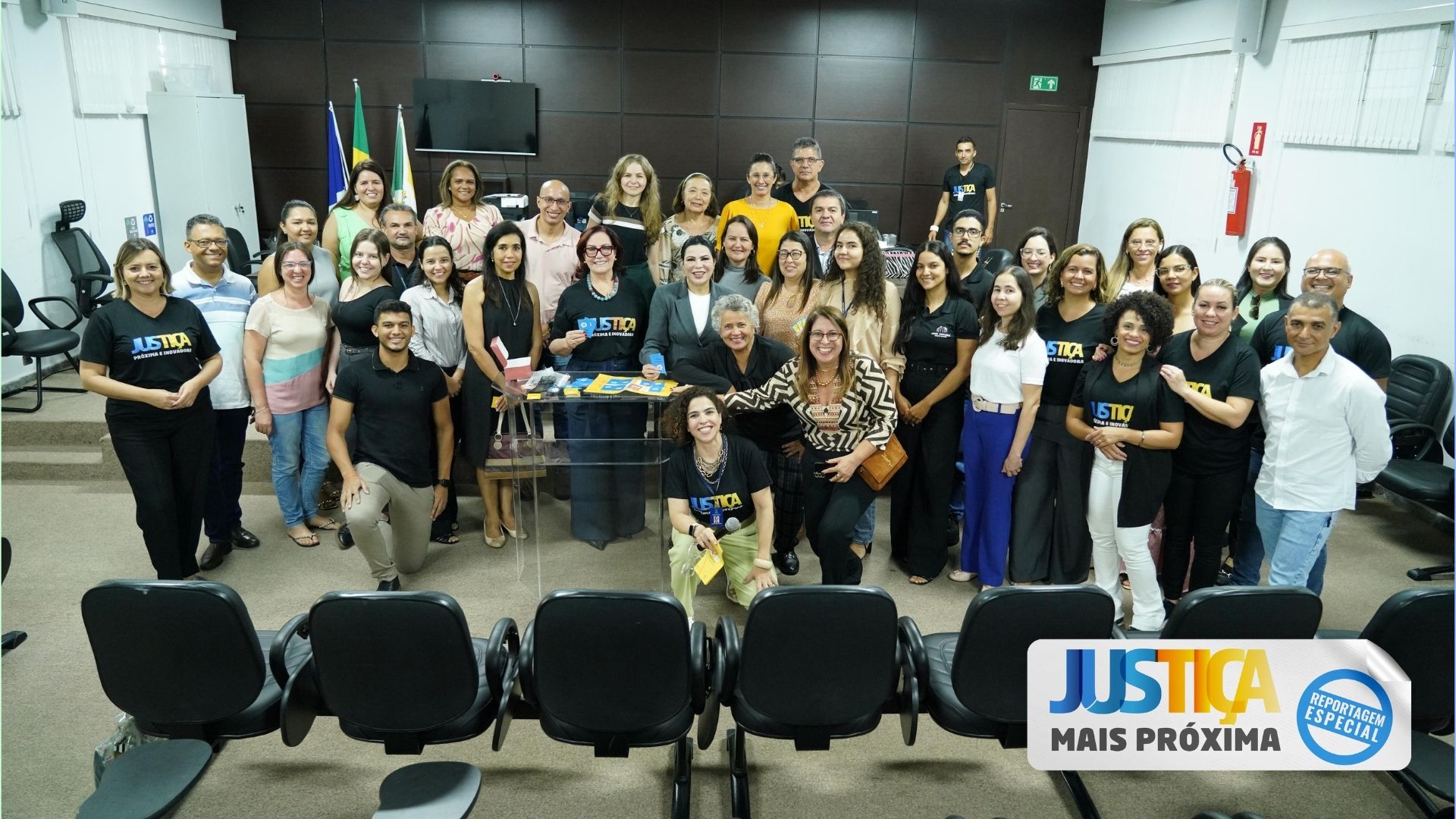 Fotografia colorida de uma sala onde um grande grupo de pessoas estão em pé posando para foto no centro de uma sala com duas paredes brancas e uma de madeira marrom ao fundo. Na frente dessas pessoas tem vários bancos pretos estofados dispostos