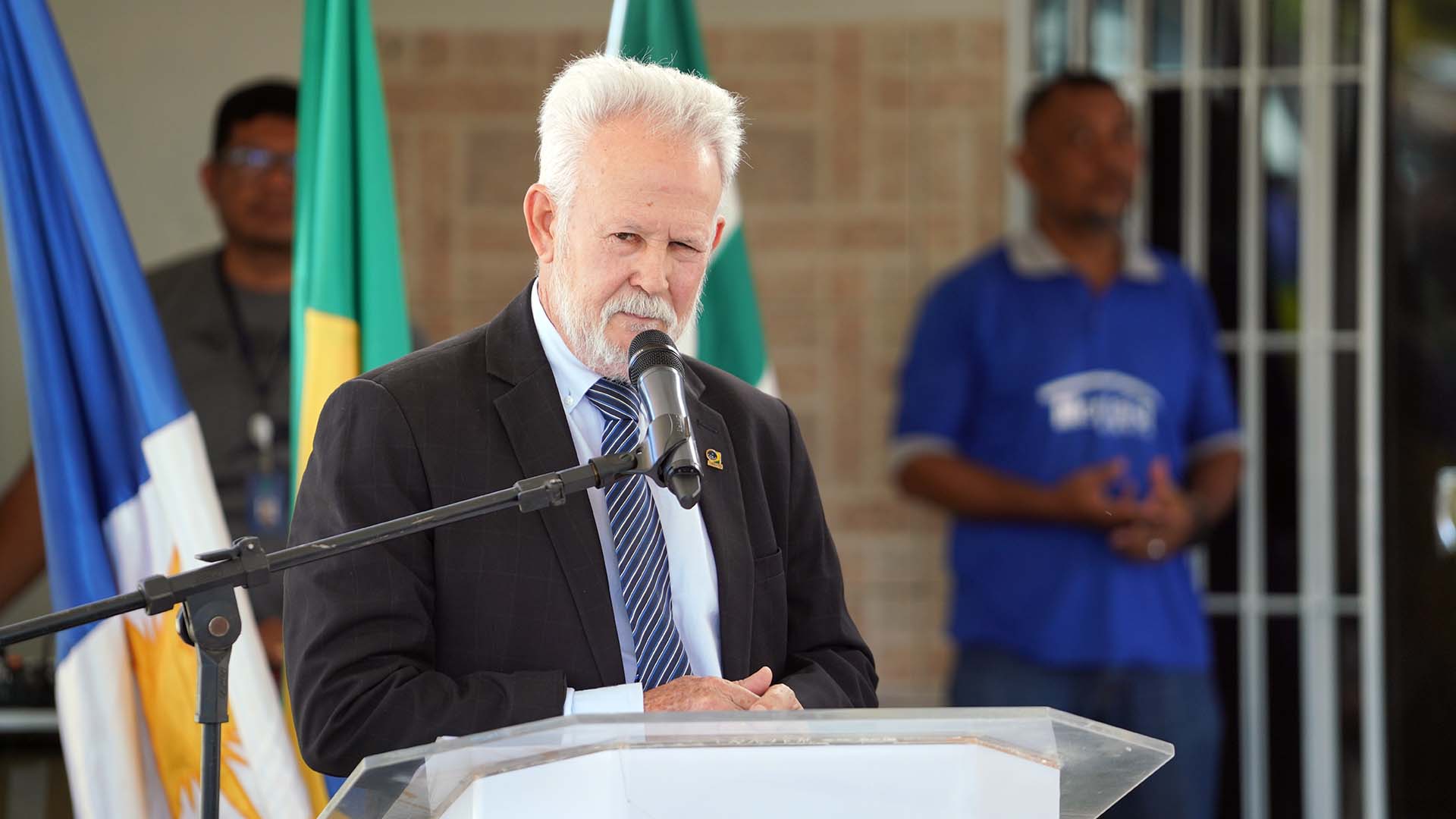 Desembargador João Rigo Guimarães, homem de cabelos e barba brancos, ele usa terno escuro, camisa branca e gravata com listras azuis, cinzas e brancas; ele fala ao microfone
