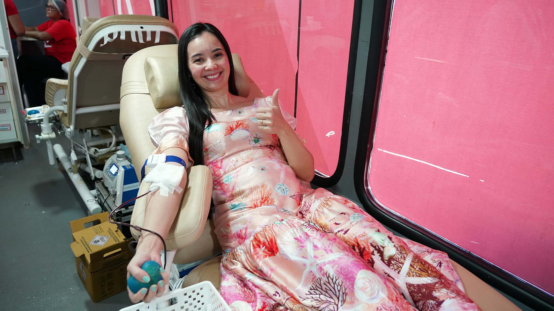 Uma doadora do sexo femino sentada, doando sangue; ela usa vestido estampando em tons de rosa e está sorrindo.