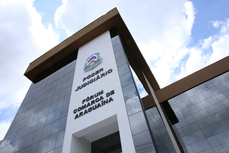 Prédio espelhado, com uma faixa de concreto bem grande e branca ao meio e dentro o símbolo da Bandeira do Tocantins e abaixo letras pretas, o teto do prédio é caramelo, como a foto foi feita de baixo pra cima, pega o céu com nuvens e um pouco de azul. 