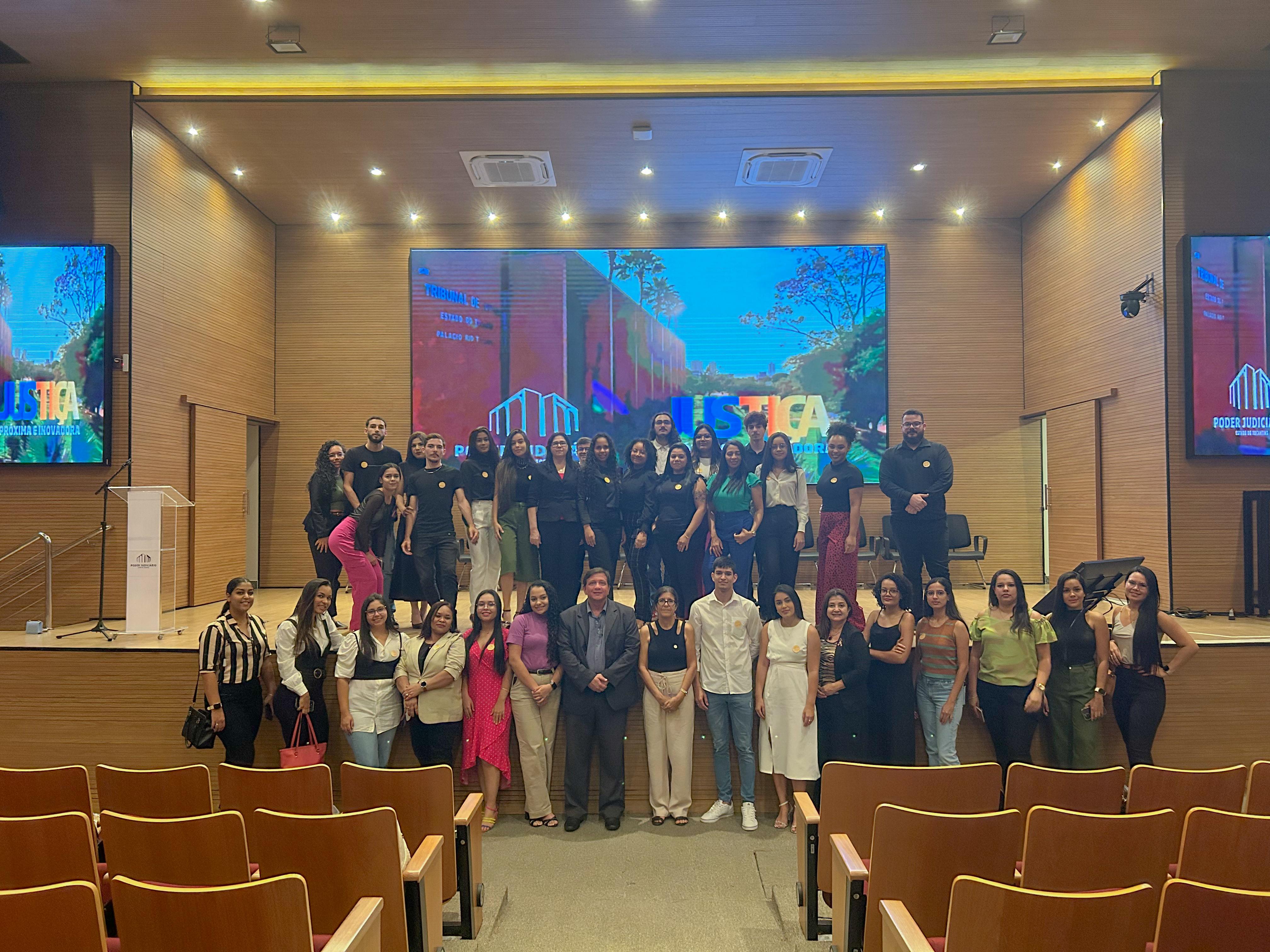 Auditório do Tribunal de Justiça do Tocantins na cor caramelo, com várias luzes no teto, no fundo tem um telão com a foto do TJTO, a frente do telão tem 35 pessoas com roupas nas cores preta, rosa, branca, listradas e verde, todas elas estão pousando para a foto, as pessoas são de diferentes faixa-etária   