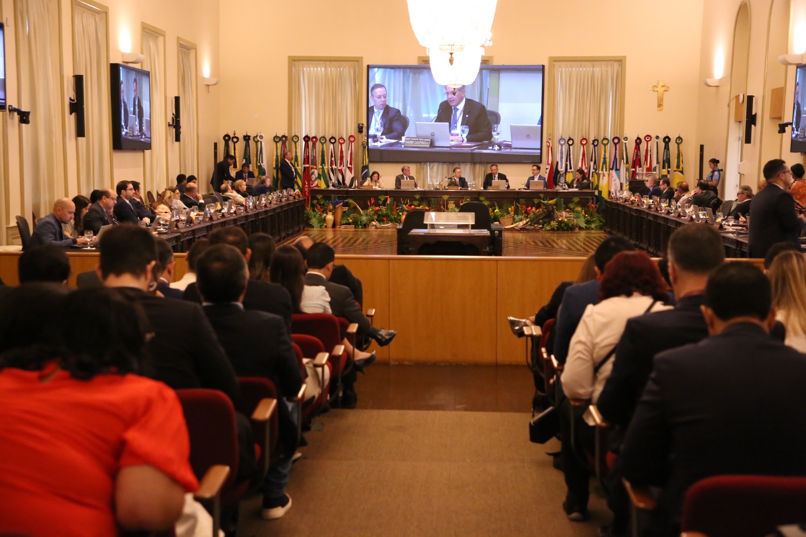 Imagem de uma grande sala com um audtório em primeiro plano, onde estão pessoas sentadas, asistindo a programação ao fundo, onde estão reunidos os desembargadores