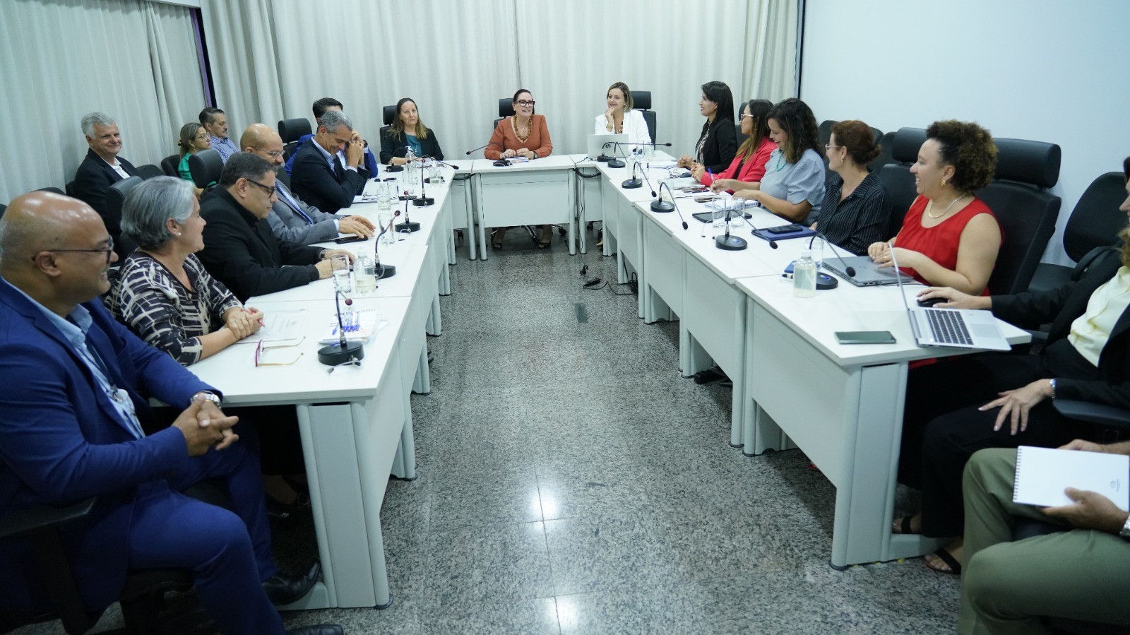 Equipes de transição reunidas numa sala, com mesas brancas em formato em U cadeiras pretas; ao fundo a atual presidente do TJTO e a presidente eleita (esq)
