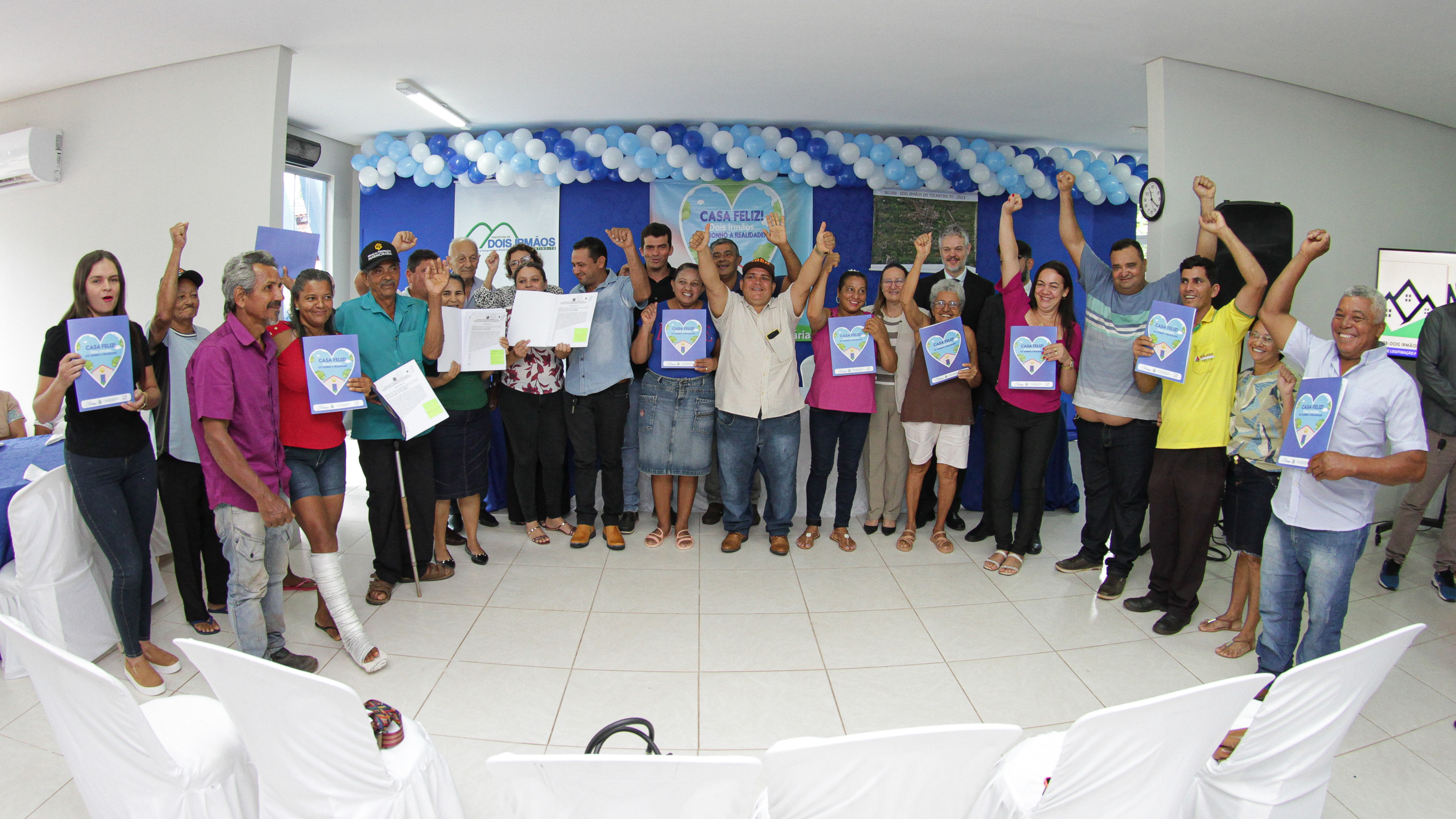 Fotografia colorida onde um grupo de pessoas comemoram com os braços para cima enquanto seguram documentos de regularização fundiária em frente a uma parede azul com balões azul claro, branco e azul escuro 