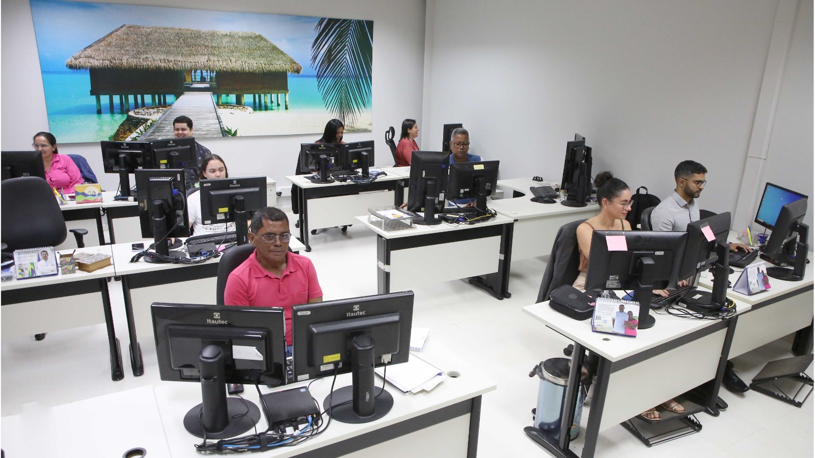 Em uma sala com mesas e computadores, nove servidores trabalham em computadores; ao fundo um quadro grande com uma imagem paradisíaca de uma cabana, um mar azul-turquesa e uma folha de coqueiro