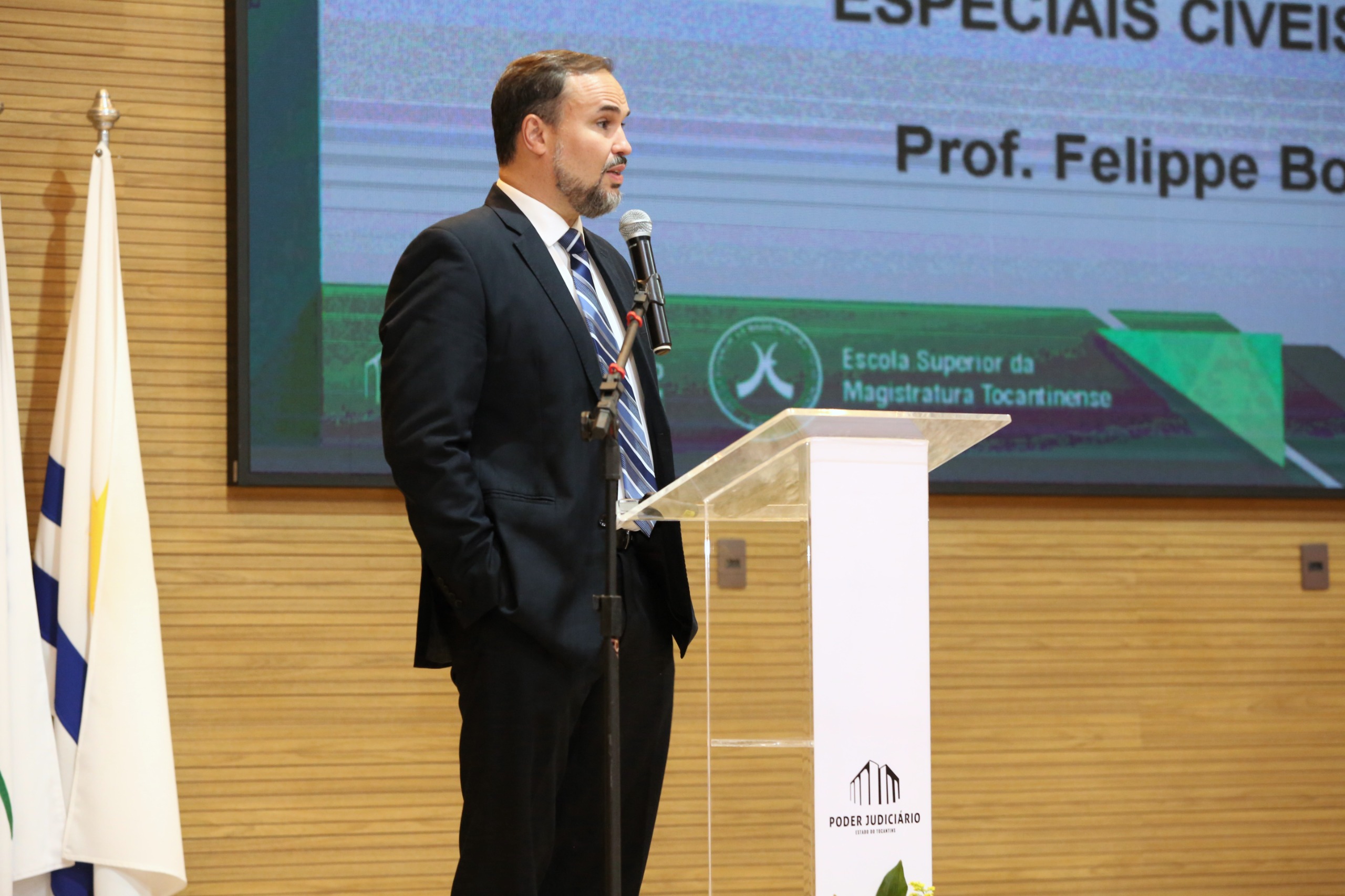 Homem branco está em pé e ao lado, sentado um outro homem de terno escuro acompanha palestra