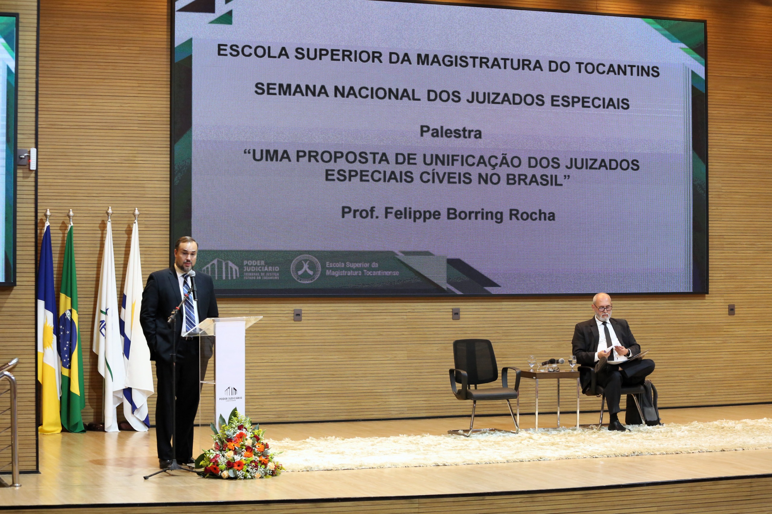 Homem branco está em pé e ao lado, sentado um outro homem de terno escuro acompanha palestra