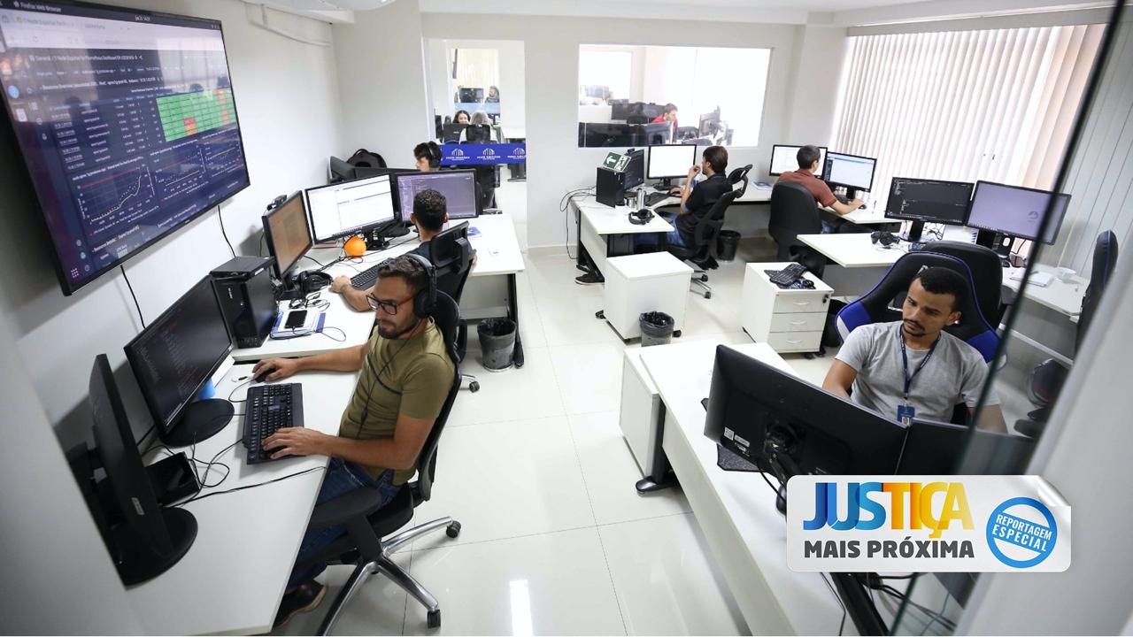 Seis servidores homens sentados, três de frente, trabalhando em computadores, e outros na sala ao fundo, vistos por janela e porta de vidro, em departamento da Diretoria de Tecnologia da Informação do TJTO; na imagem aparecem mesas e móveis brancos, paredes e persiana brancas e um monitor na parede, à esquerda.