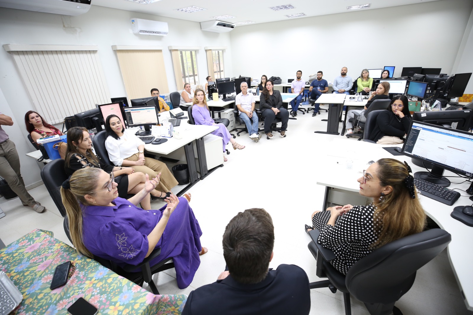 quatro juízes (um homem e três mulheres) conversando com servidores que trabalharão no bloco de competência do Sistema de Juizados Especiais; eles estão sentados, em uma sala de trabalho; na foto aparecem mesas brancas, cadeiras pretas e vários monitores sobre as mesas, além de ar-condicionados e persianas nas janelas 