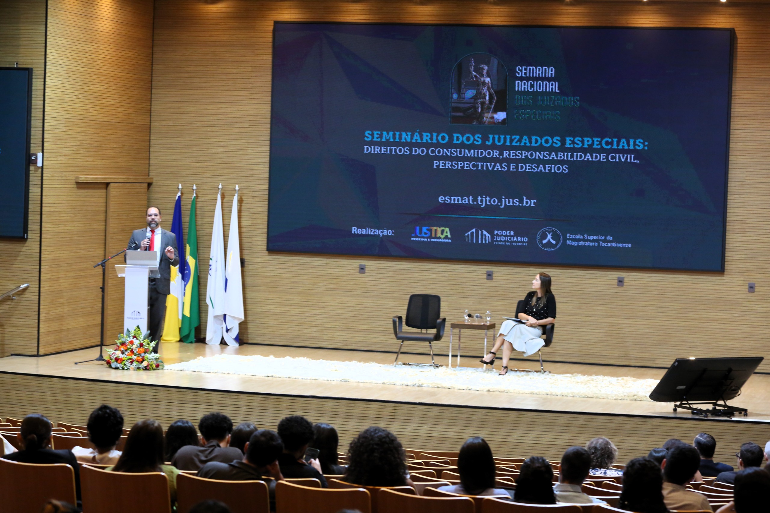 Homem de terno atrás do púlpito fala para uma plateia; no palco uma mulher está sentada