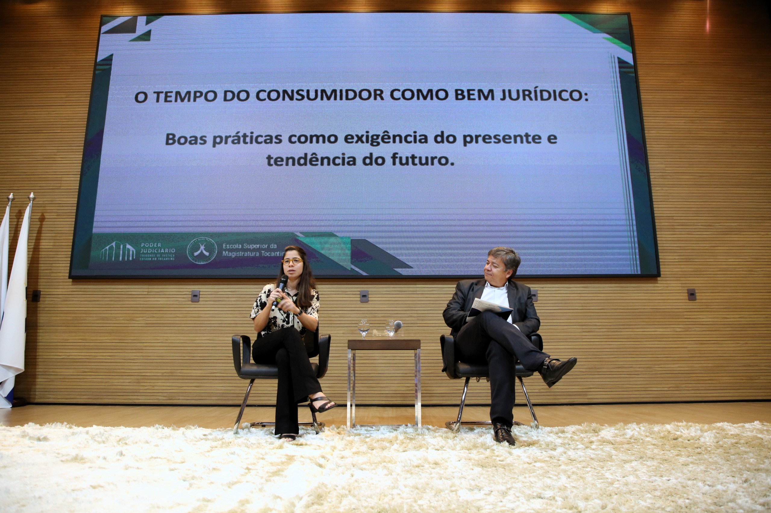 Homem de terno atrás do púlpito fala para uma plateia; no palco uma mulher está sentada