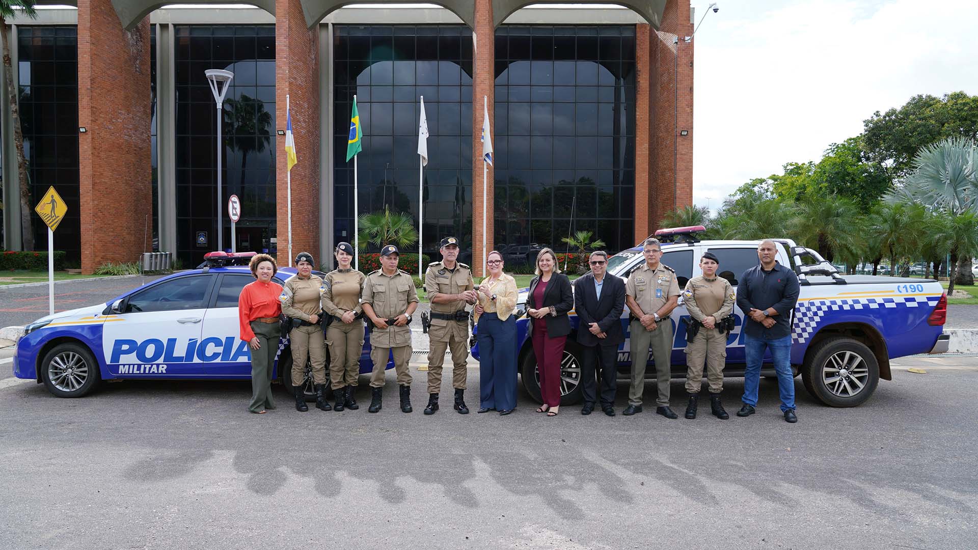 Presidente do TJTO ao centro da imagem de calça azul e camisa amarela, ao lado da diretora geral, de roupa berinjela e do diretor administrativo de terno escuro. Eles estão entre policiais militares, na frente de duas viaturas da PM, em frente a sede do Tribunal