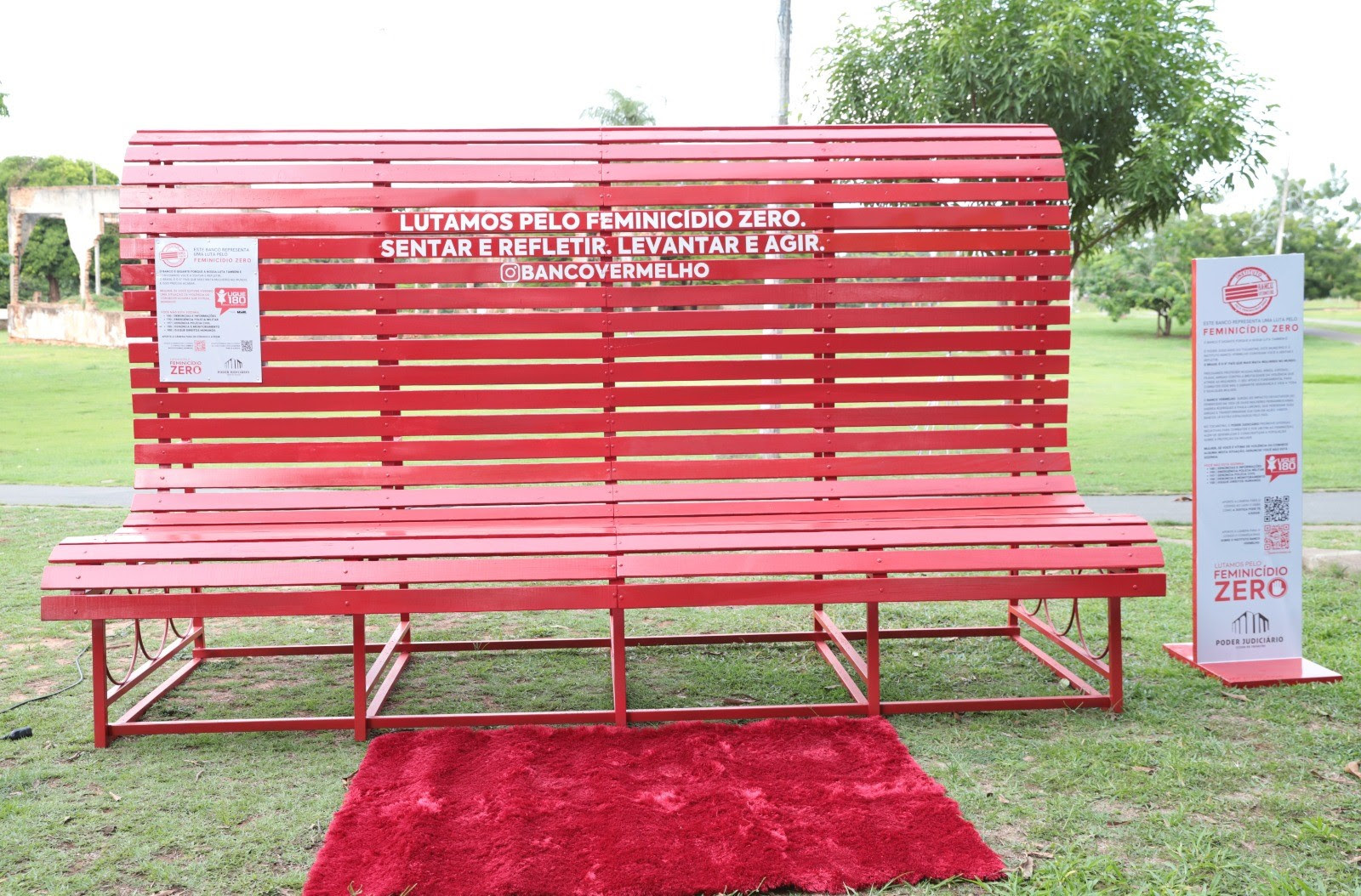 Fotografia colorida de um grande banco vermelho disposto sobre a grama verde em um espaço aberto a luz do dia. Em sua frente, está um pequeno tapete também vermelho. 