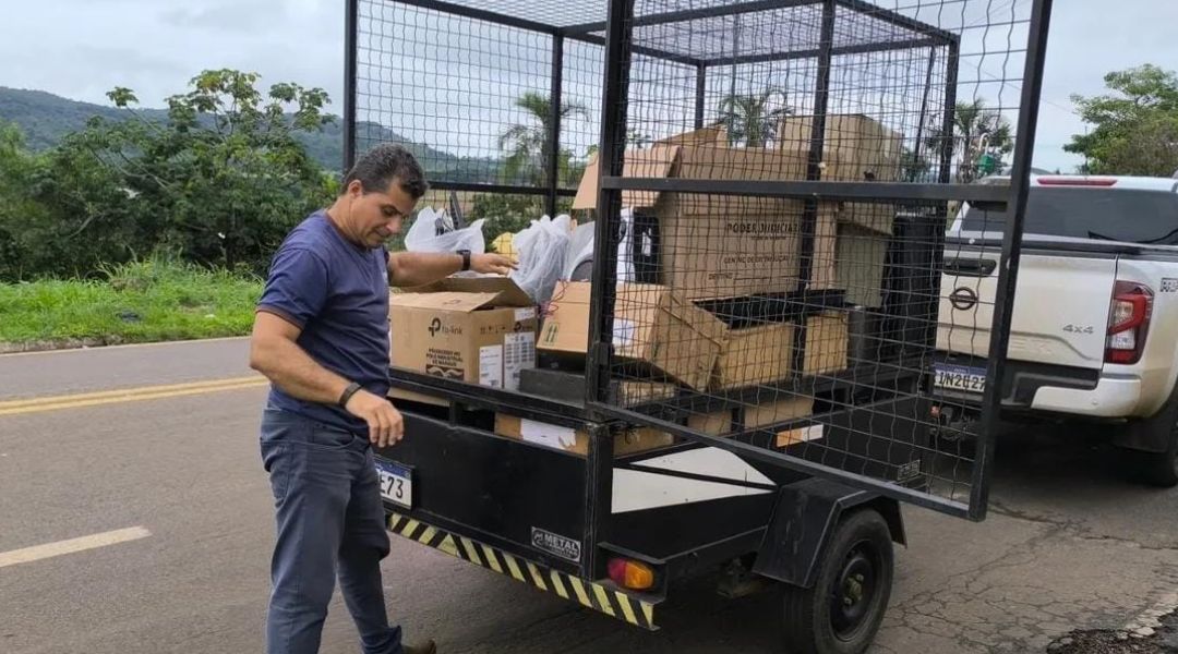  Fotografia colorida onde um homem de camisa azul escuro e calça preta está colocando lixo em uma gaiola móvel que está encaixada em uma caminhonete. Ele está em uma pista aberta 