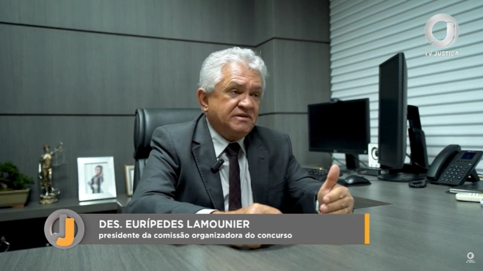 Homem idoso de cabelos grisalhos trajando terno cinza escuro, gesticula enquanto fala sentado à mesa de escritório com computador com duas telas. Ao fundo, parede revestida em madeira acinzentada e janela com persiana bege