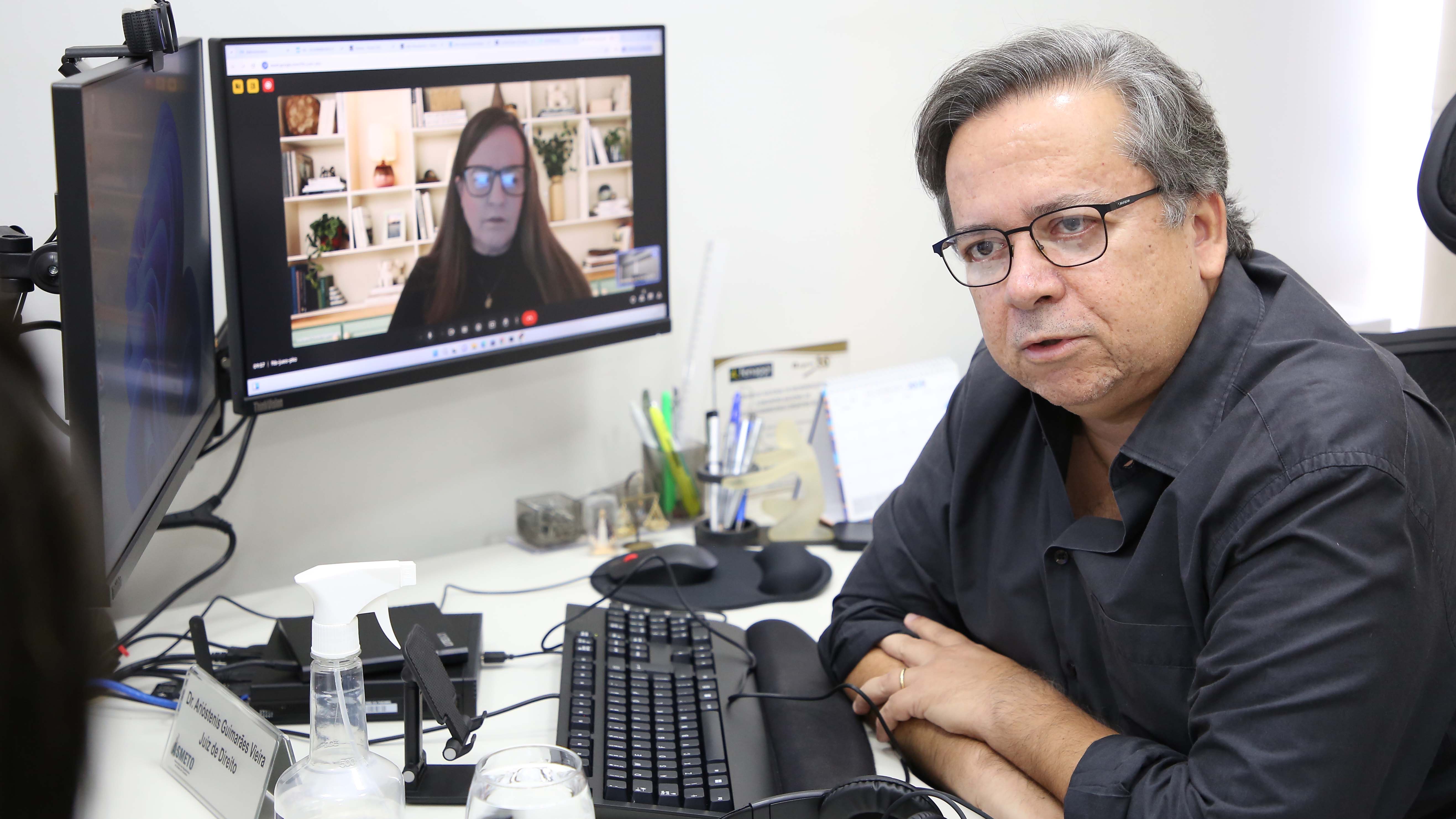 Homem de cabelos lisos e grisalhos, de terno preto, sentado de braços sobre mesa de trabalho em frente a computador de duas telas durante reunião virtual com mulher de cabelos longos e liso, de óculos de grau