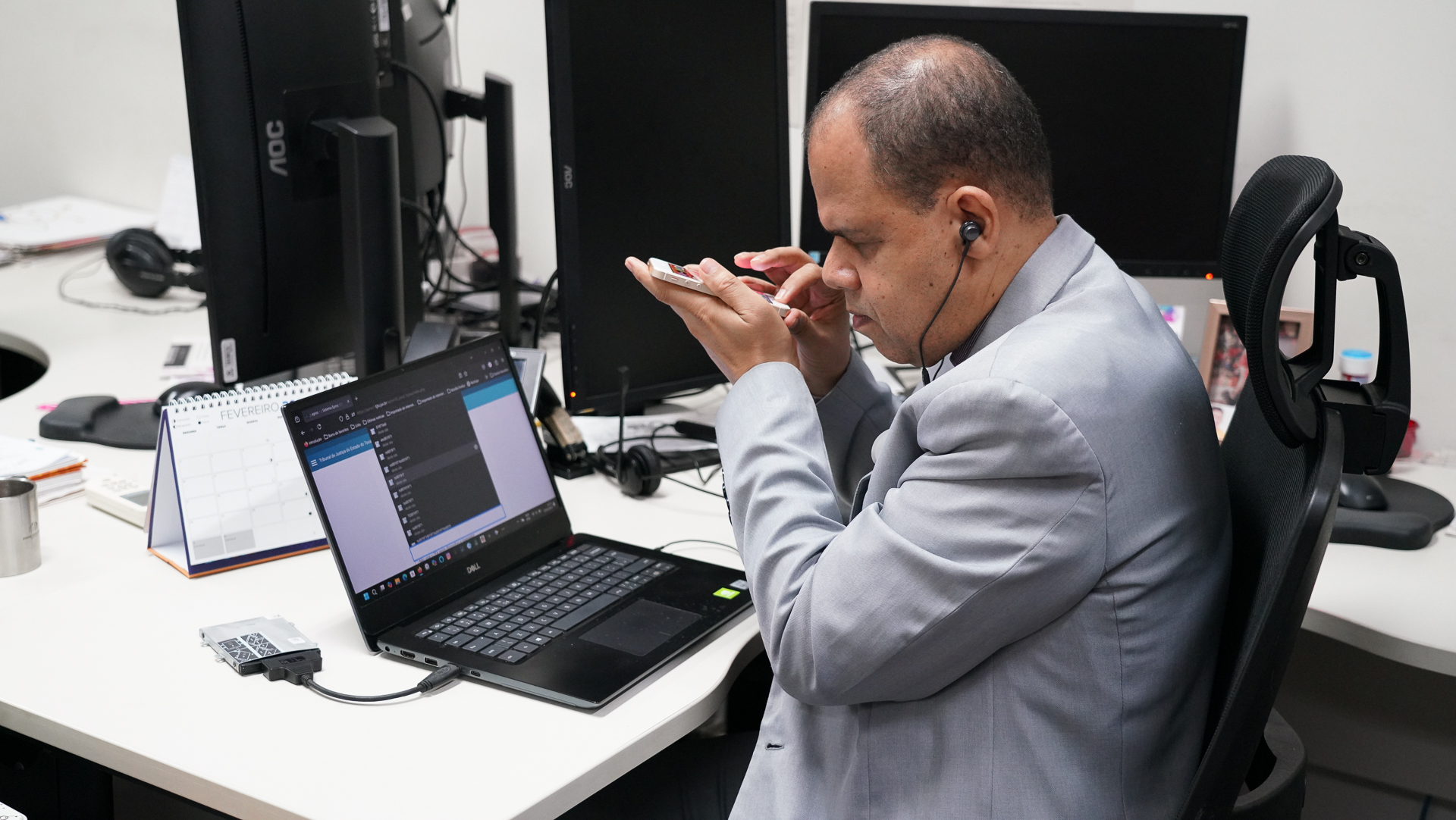 Homem de terno sentado em uma mesa, em frente ao computador. Ele é cego e está com um celular na mão e fone de ouvido