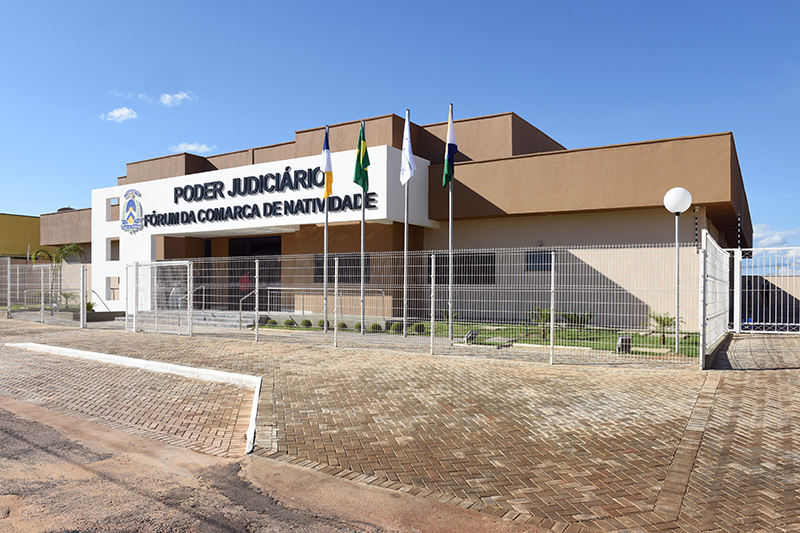 Fotografia colorida da fachada do Fórum da Comarca de Natividade. Em frente e ao redor da Comarca, existe uma grade de proteção branca, e o chão é composto por tijolos de padrões iguais na calçada. Ao fundo, um belo céu azul está projetado 