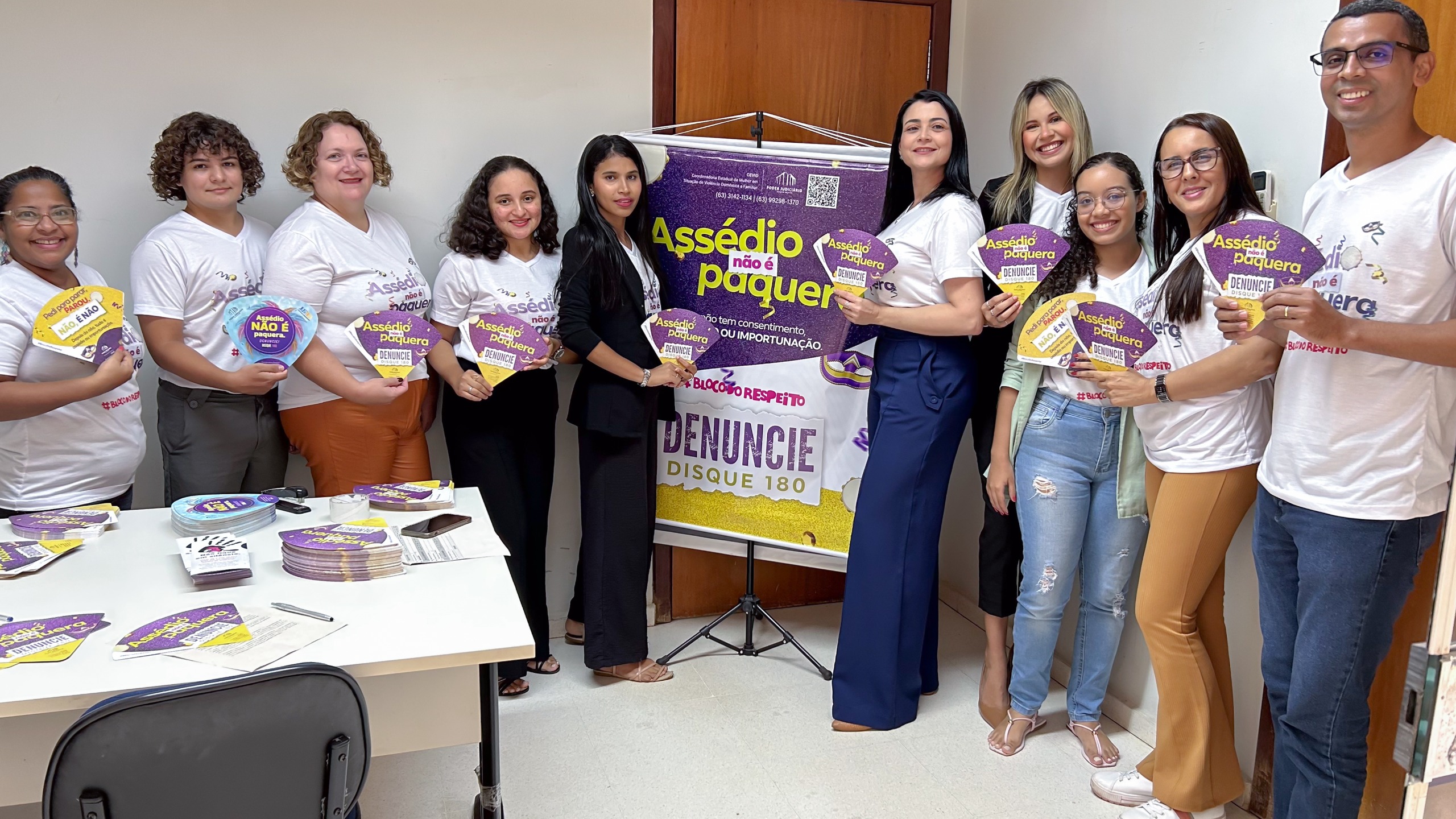 Na imagem tem um grupo de dez pessoas, composto por homens e mulheres, está reunido em uma sala, segurando materiais de campanha sobre o combate ao assédio no Carnaval. A maioria veste camisetas brancas com a frase "Assédio não é paquera". No centro da imagem, há um banner roxo e amarelo com a mesma mensagem, destacando o canal de denúncia "Disque 180". Tem uma mesa com materiais da campanha, como panfletos, leques e adesivos. Todos sorriem e olham para a câmera, demonstrando engajamento na iniciativa.
