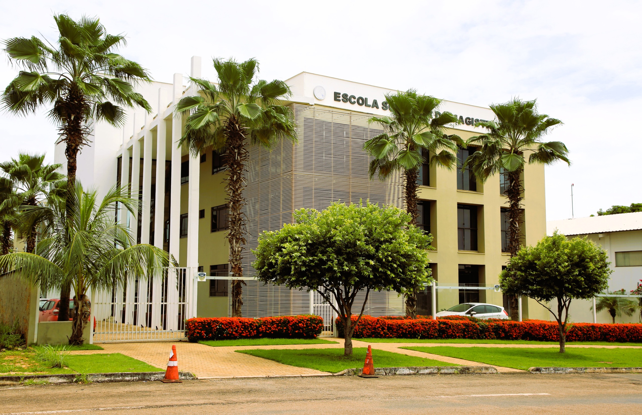 Na imagem aparece o prédio da Escola Superior da Magistratura Tocantinense, na cor amarelo e bege. O prédio é cercado por uma área bem cuidada com gramados, arbustos e árvores, incluindo palmeiras. 