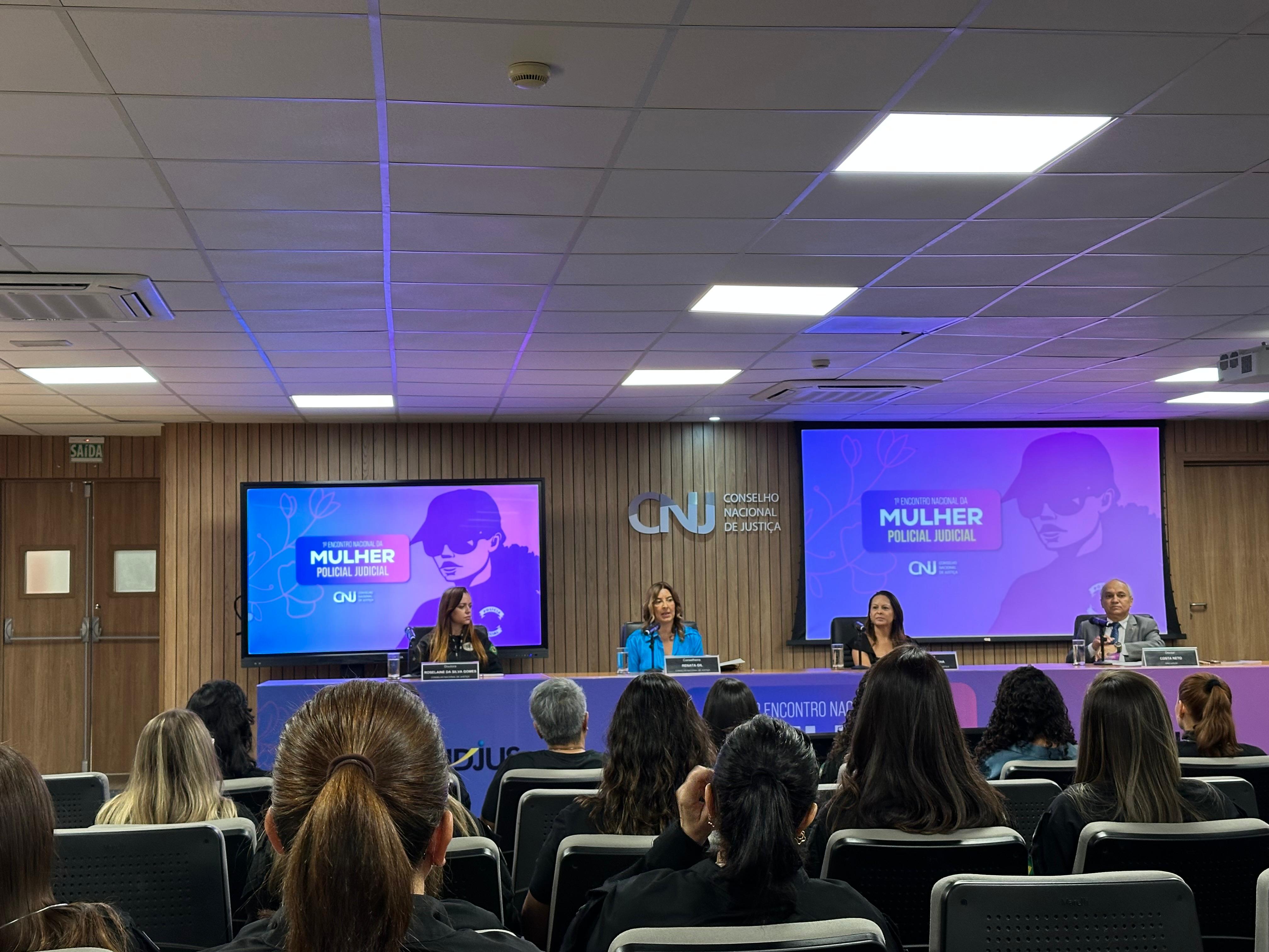 A imagem mostra várias mulheres e um homem em um auditório, durante o primeiro Encontro Nacional da Mulher Policial Judicial. Sendo que na composição da imagem tem três mulheres e um homem, palestrantes, sentados de frente para um grande número de outras pessoas que também estão sentadas participando do evento. Ao fundo há um telão com o nome do encontro. 