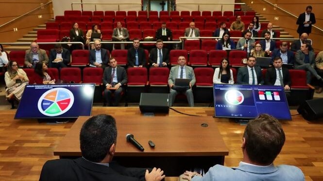 A imagem mostra um auditório com várias pessoas sentadas em fileiras, direcionadas para um palco com uma mesa, onde tem dois homens que aparecem de costa pra imagem e de frente pra plateia.  Na cena também aparecem duas televisões exibindo dados de uma apresentação.