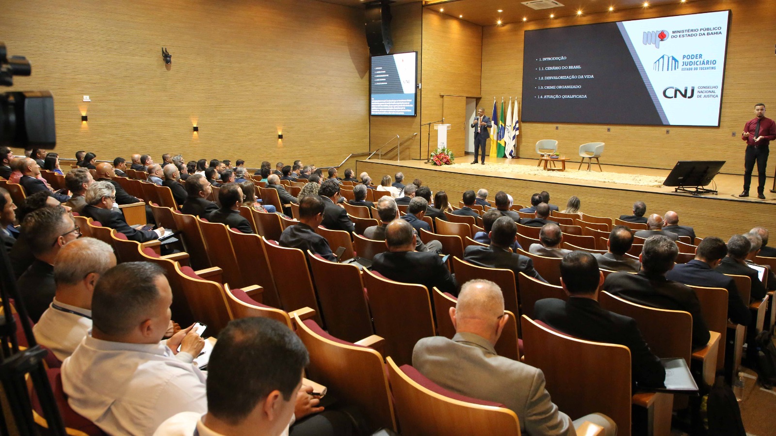 Auditório com paredes revestidas em madeira, com público sentado de costas. Ao fundo, homem fala à esquerda, e outro homem traduz em Libras. Um telão transmite informações sobre evento de Inteligência institucional
