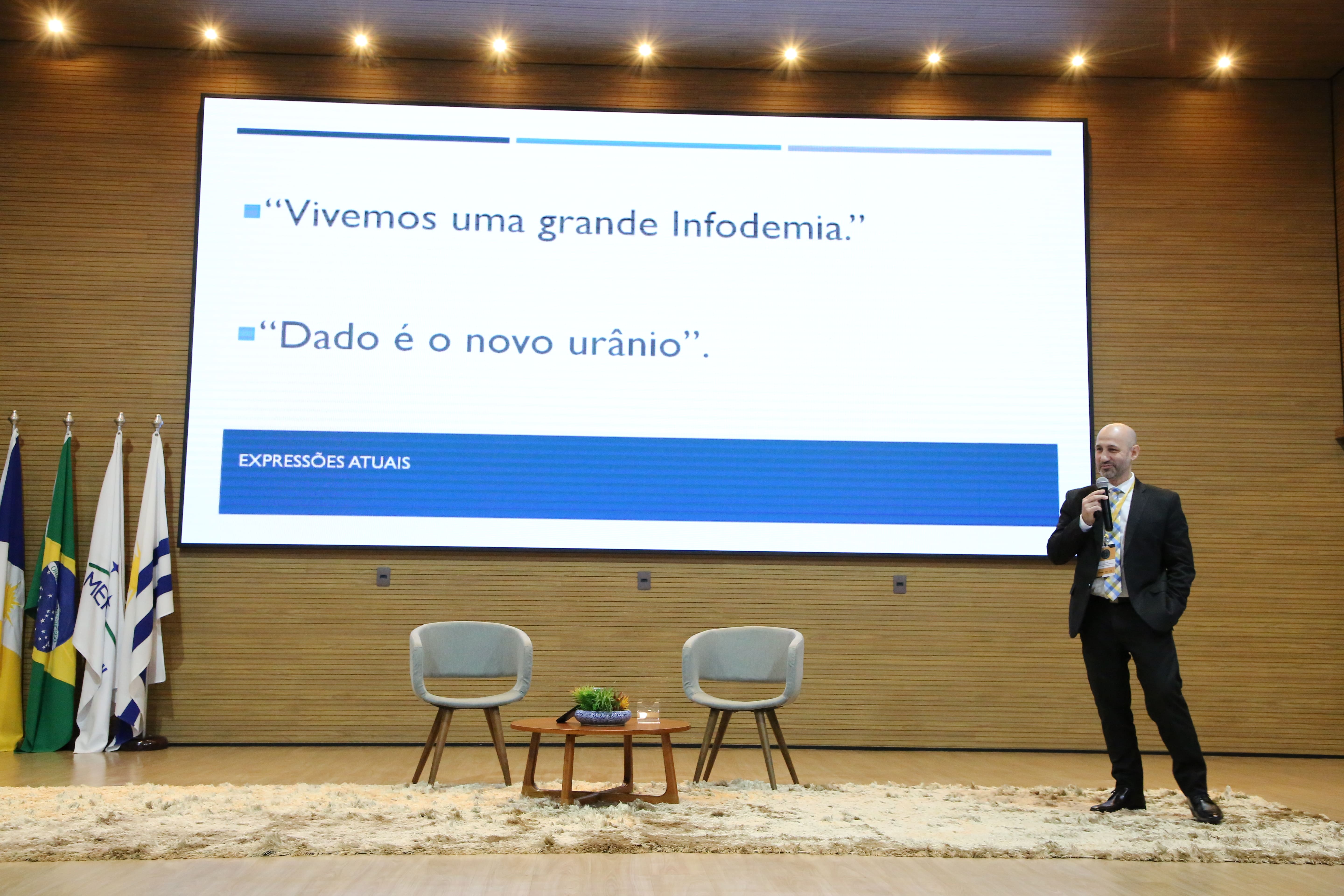 Imagem mostra o palestrante, de terno, no palco do auditório, dando uma palestra, com um projetor exibindo texto em português. O palestrante ocupa o centro da imagem, posicionado em frente a um telão com informações sobre o tema.  O fundo é neutro, composto por paredes de madeira clara e um tapete no chão.