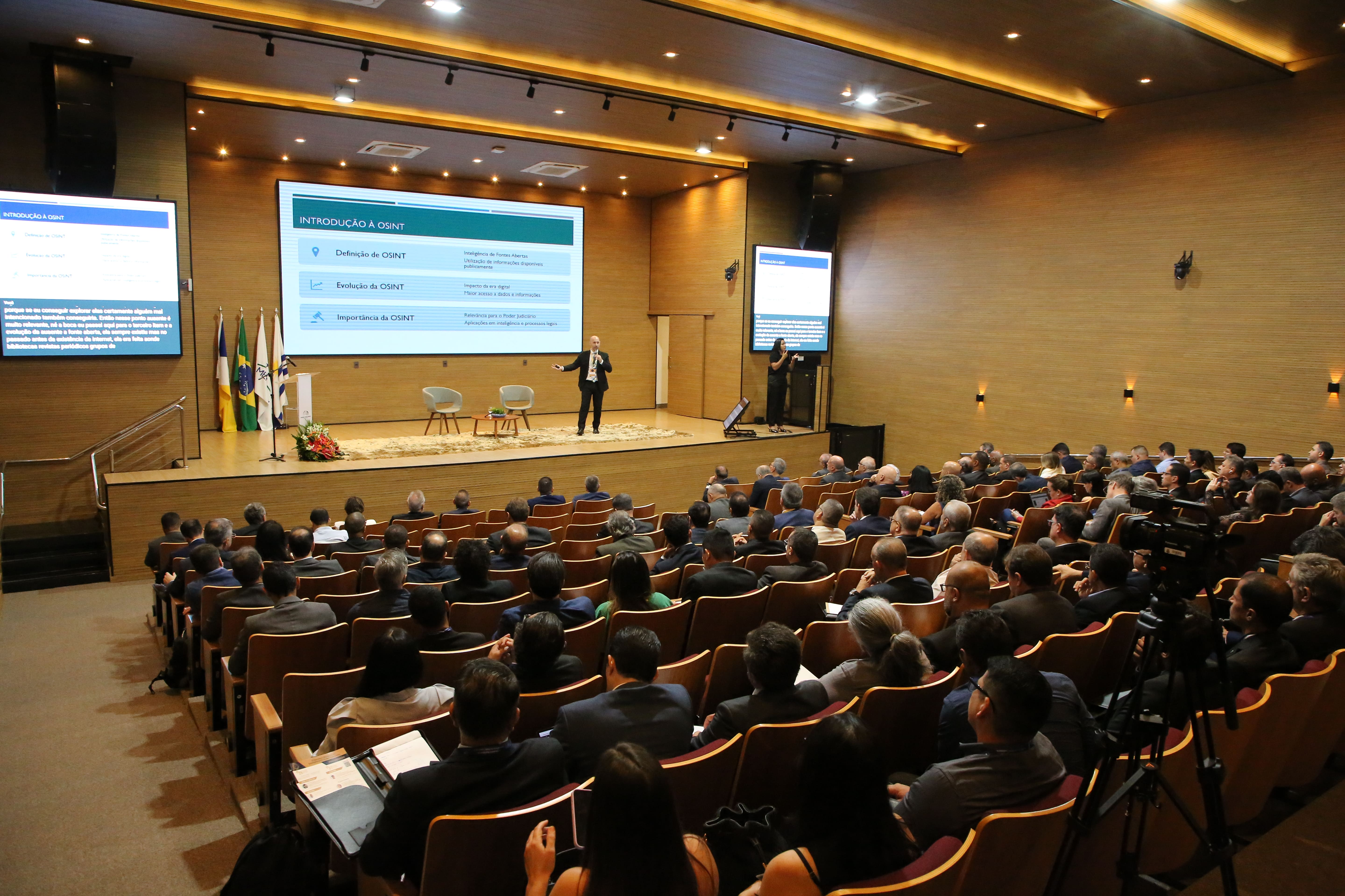 Imagem mostra um amplo auditório com muitos participantes assistindo a uma palestra.  Há várias telas grandes exibindo informações na área do palco. O espaço é bem iluminado e as pessoas estão em postura atenta, demonstrando interesse pela palestra. 